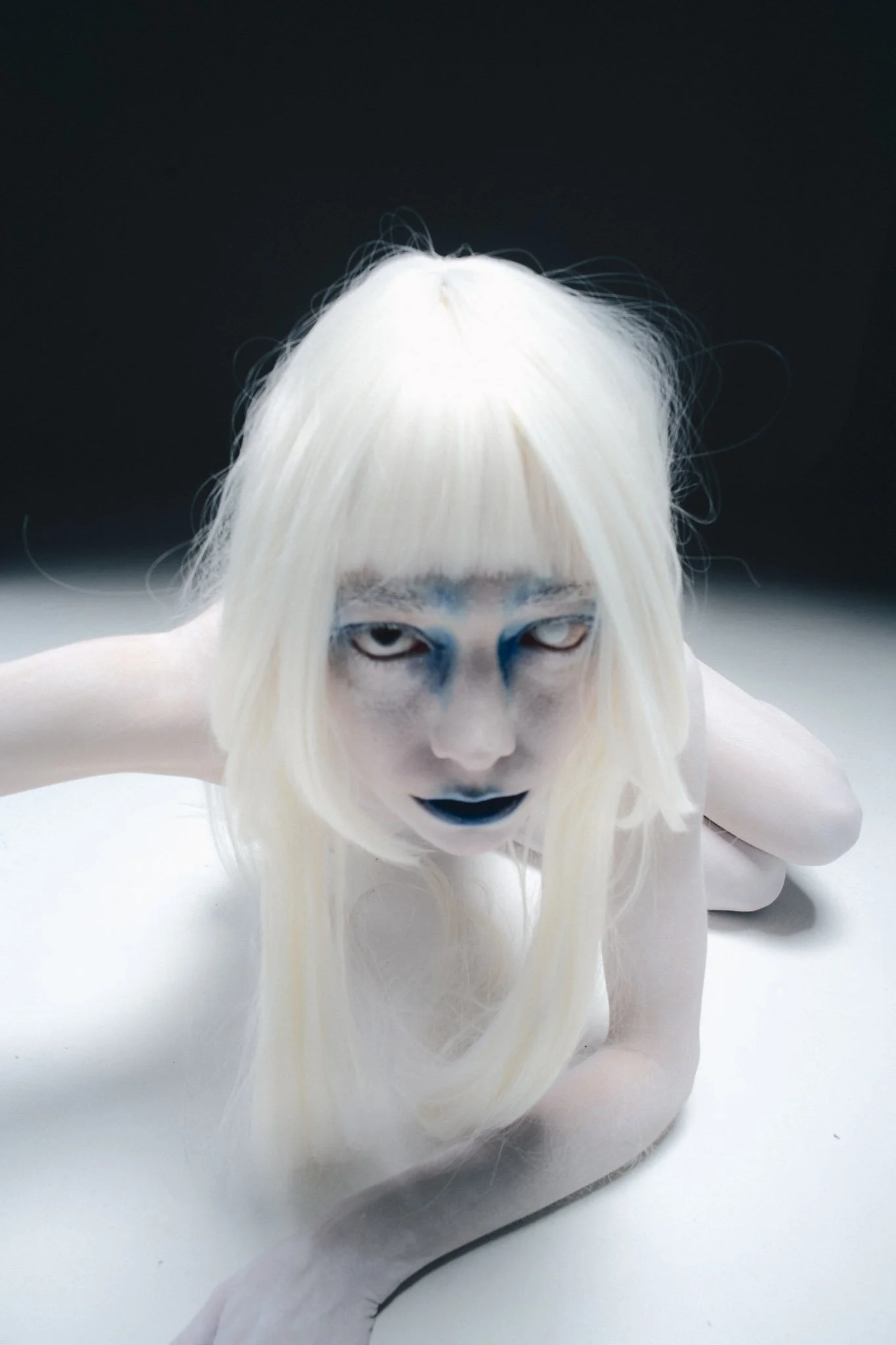 Close-up of a white doll with long platinum blonde hair and painted facial features, lying on a white surface with a dark background.