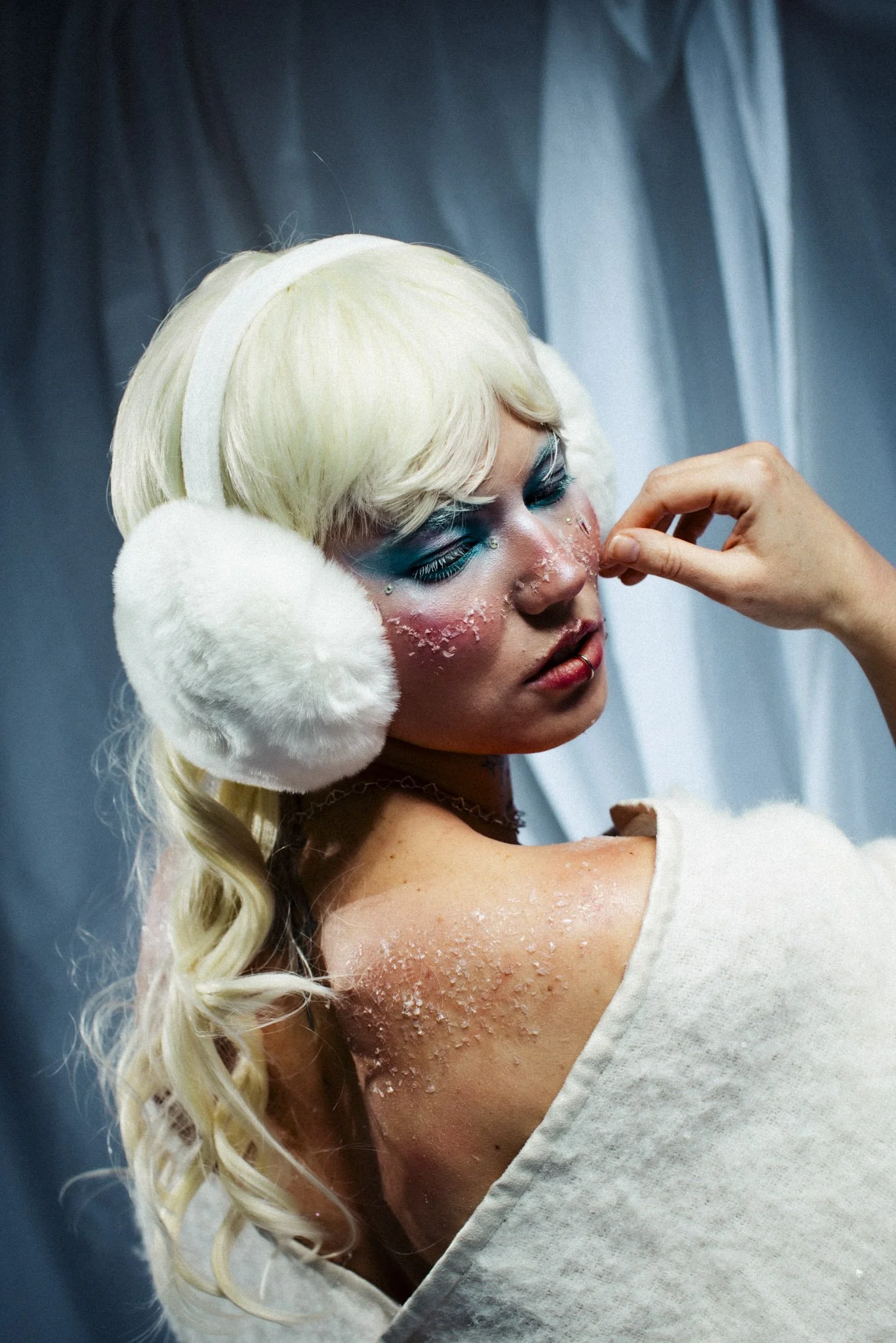 A woman with platinum blonde hair and dramatic makeup, including blue eyeshadow and rhinestones near her eyes, is having a skincare treatment applied to her face. She is wearing white, fluffy earmuffs and a white towel wrapped around her shoulders, w