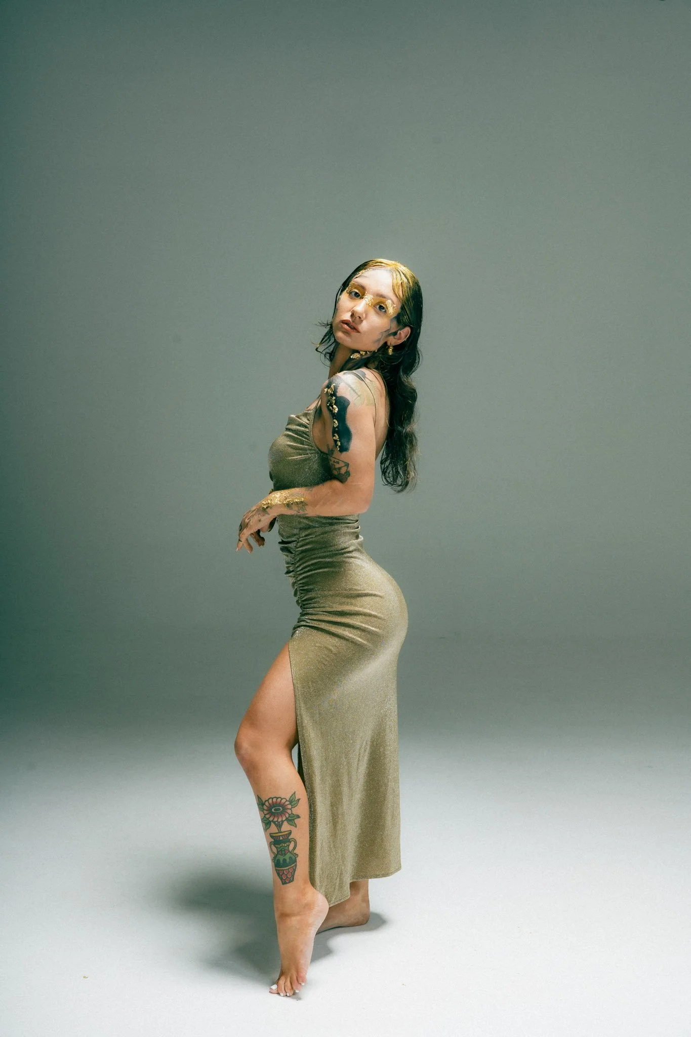 A woman with dark, wavy hair and tattoos on her arms and leg, wearing a shiny gold dress with a high slit, poses with her head tilted and one leg slightly forward against a plain muted background.