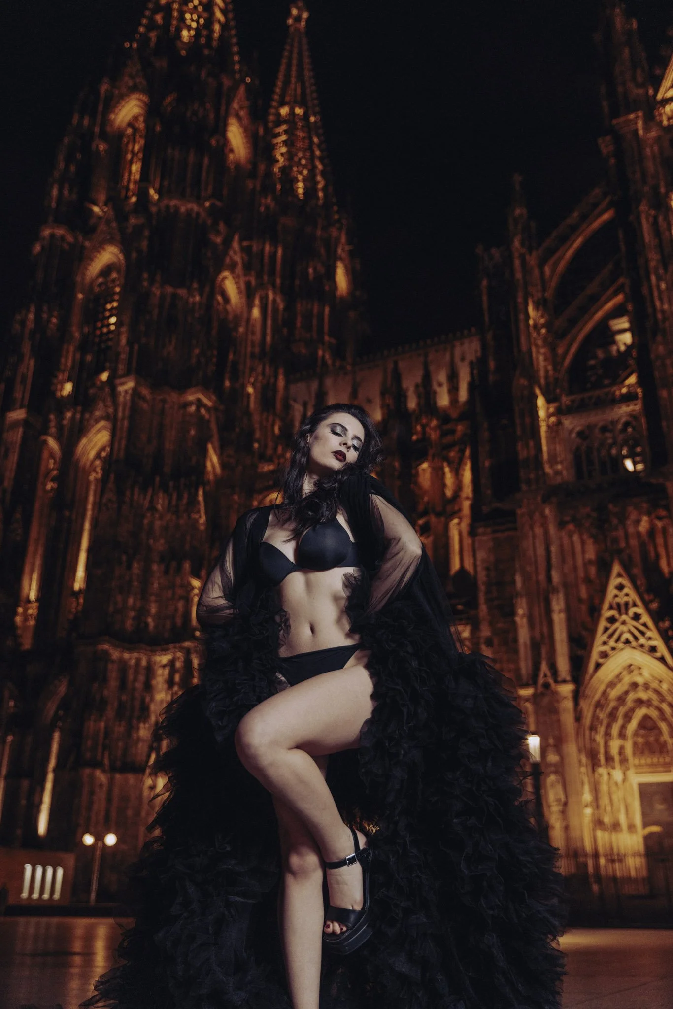 A woman in black lingerie and a black feathered skirt poses sensually in front of a gothic cathedral lit up at night.