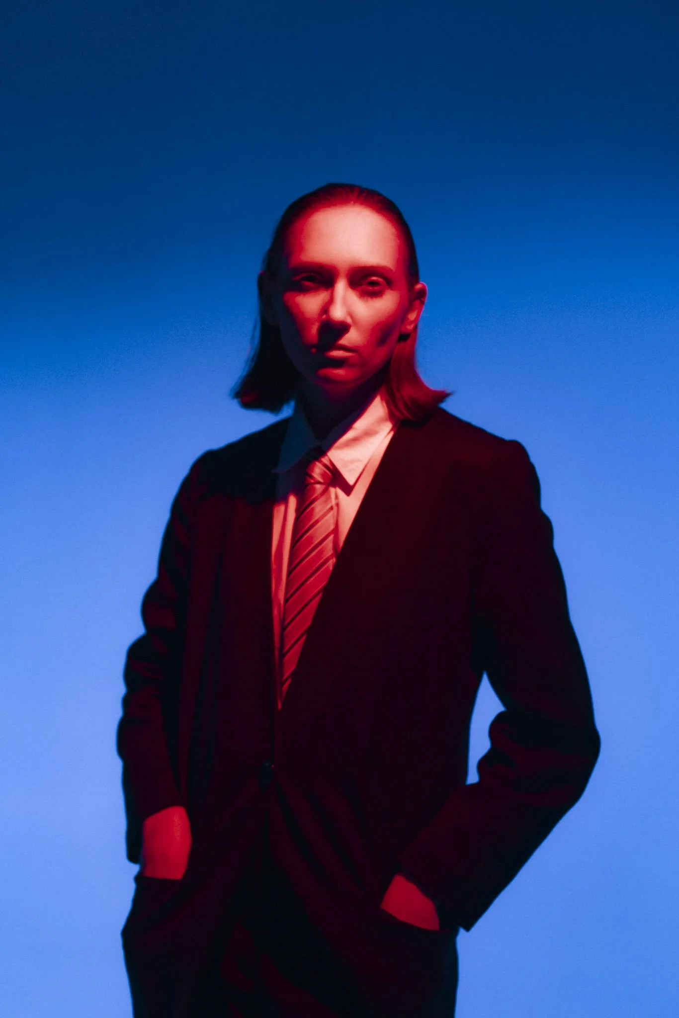 A person with long hair wearing a suit and tie, standing against a gradient blue background with red lighting on their face.