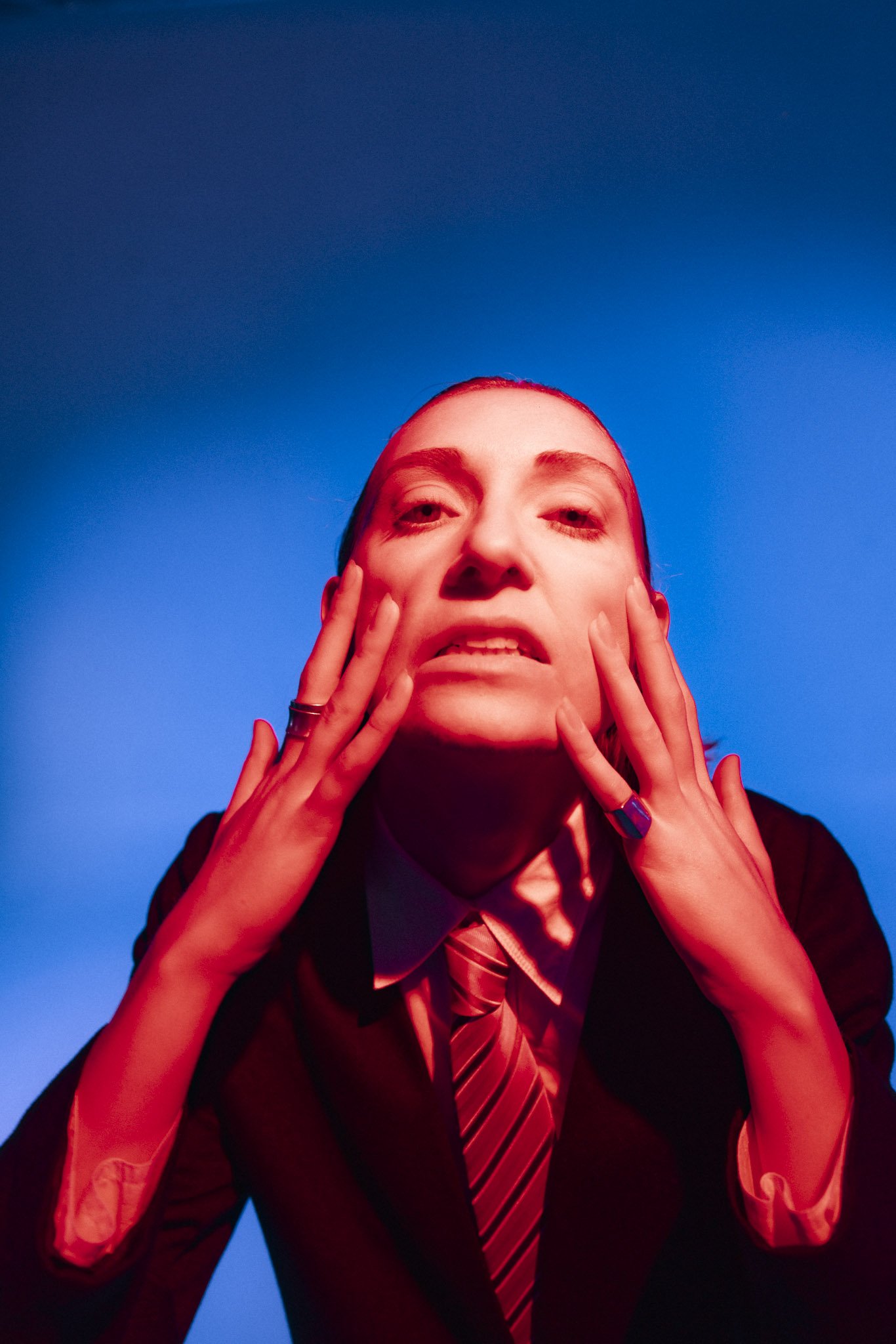 A person wearing a suit and tie, with hands on their face, taken against a background that is half red and half blue, with the person's face illuminated by the red light.