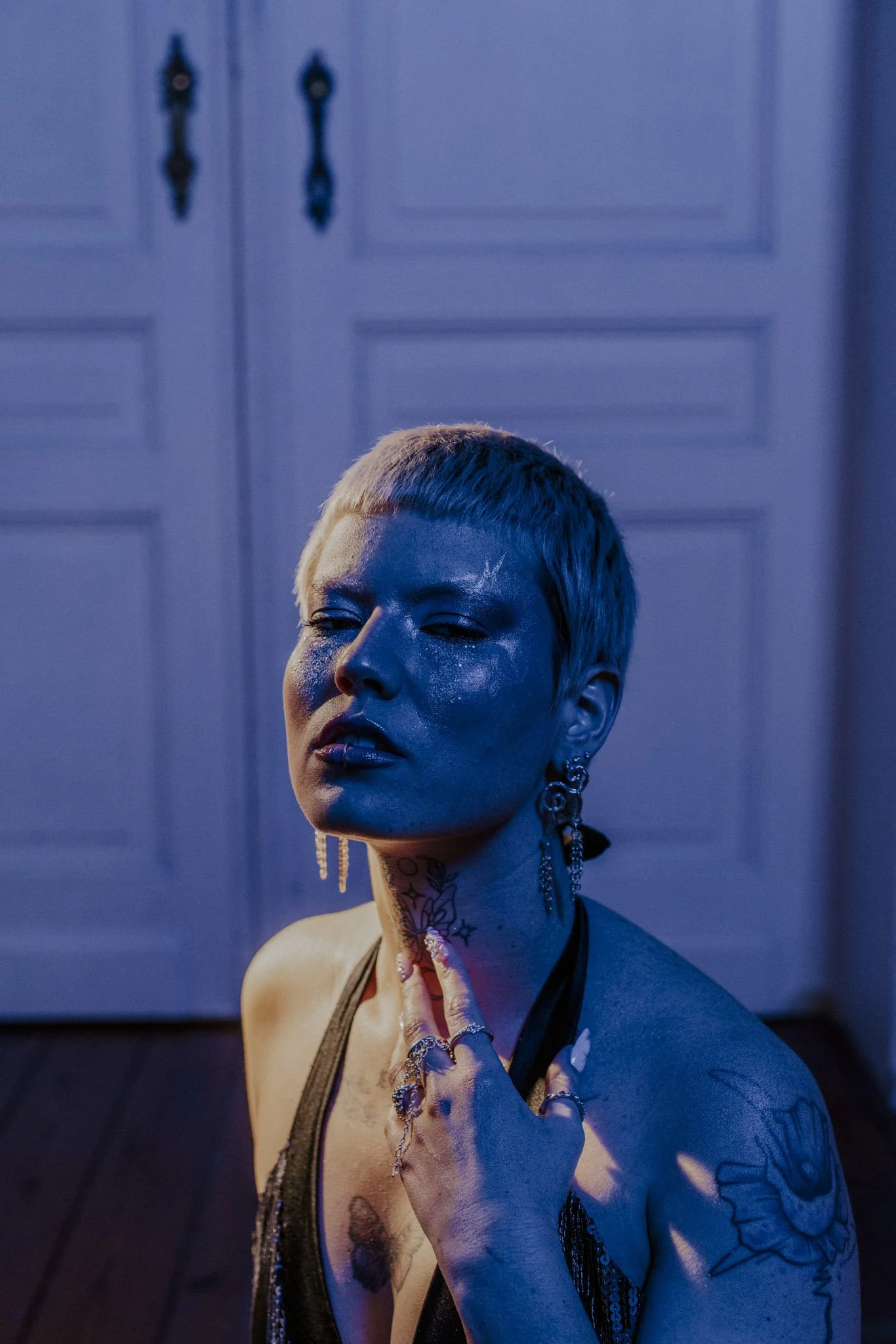 A person with short blonde hair and tattoos on their arm, neck, and chest, wearing multiple rings and jewelry, in a dimly lit room with blue and purple lighting, and a blue wardrobe in the background.