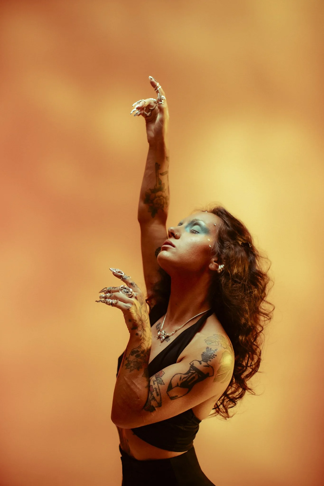 A woman with tattoos on her arms and chest, makeup with turquoise eyeshadow, and jewelry, posing with her arm extended upward against a warm orange background.