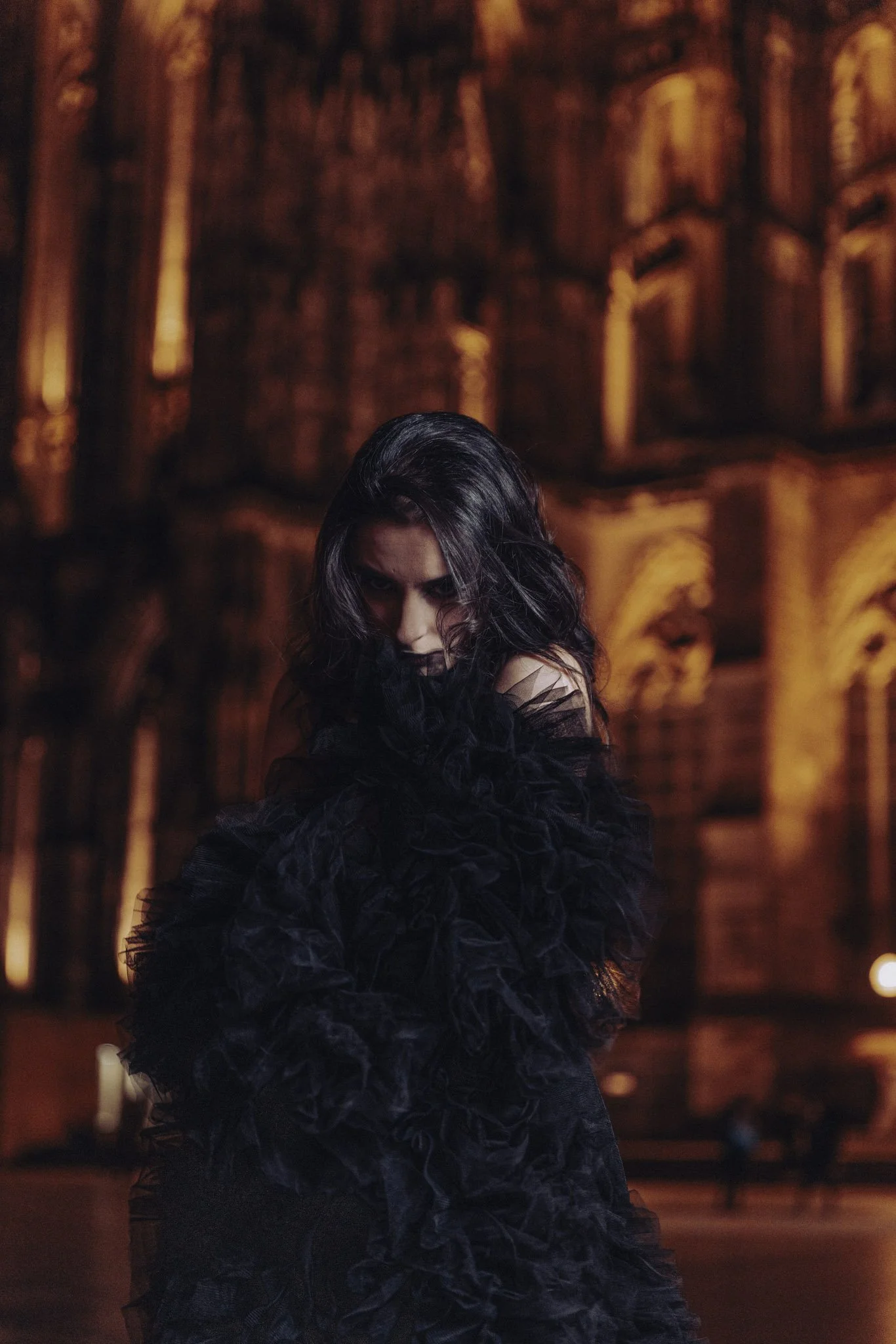 A woman with dark hair wearing a black, feathered dress poses in front of a warmly lit, ornate wooden wall with arches.