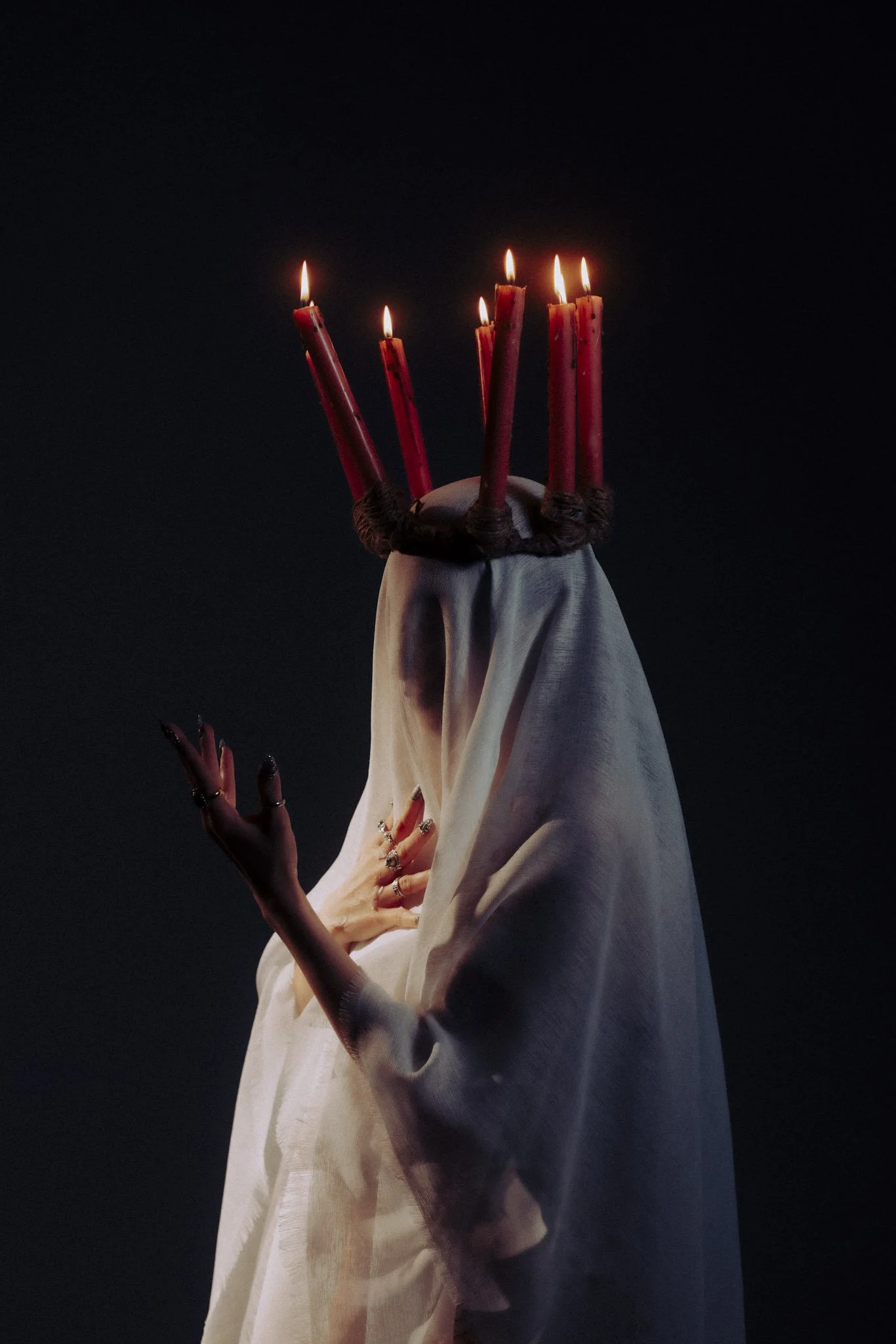 A person with a white cloth dress has seven red candles on their head, lit and burning, against a dark background.