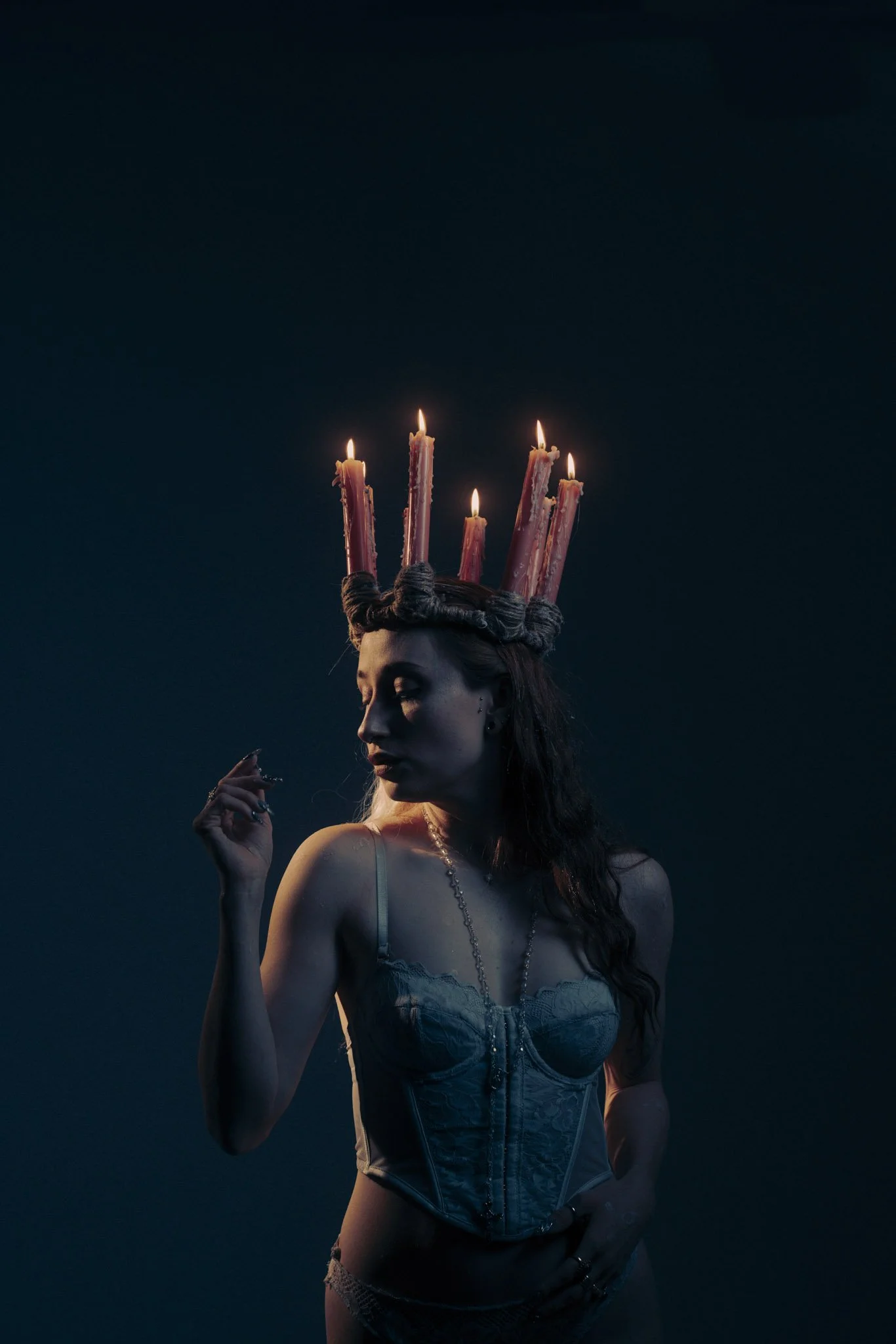 A woman with long, wavy hair wearing a lace corset and lace underwear, holding a cigarette, with a crown of lit pink candles on her head, against a dark background.