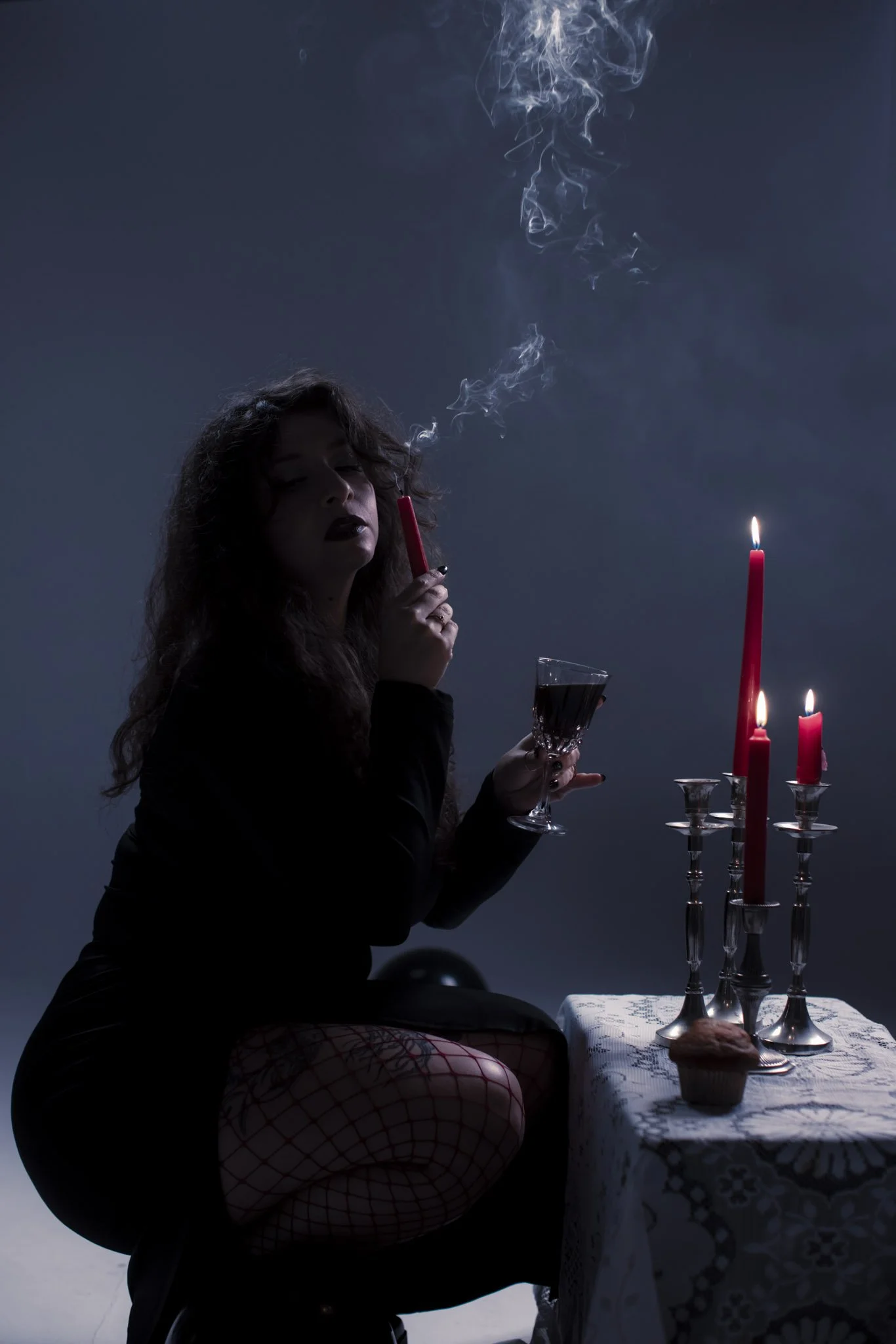 A woman with dark hair and makeup sitting at a table with lit red candles, smoking from a red cigarette holder, holding a wine glass, in a dark setting with a blue background.