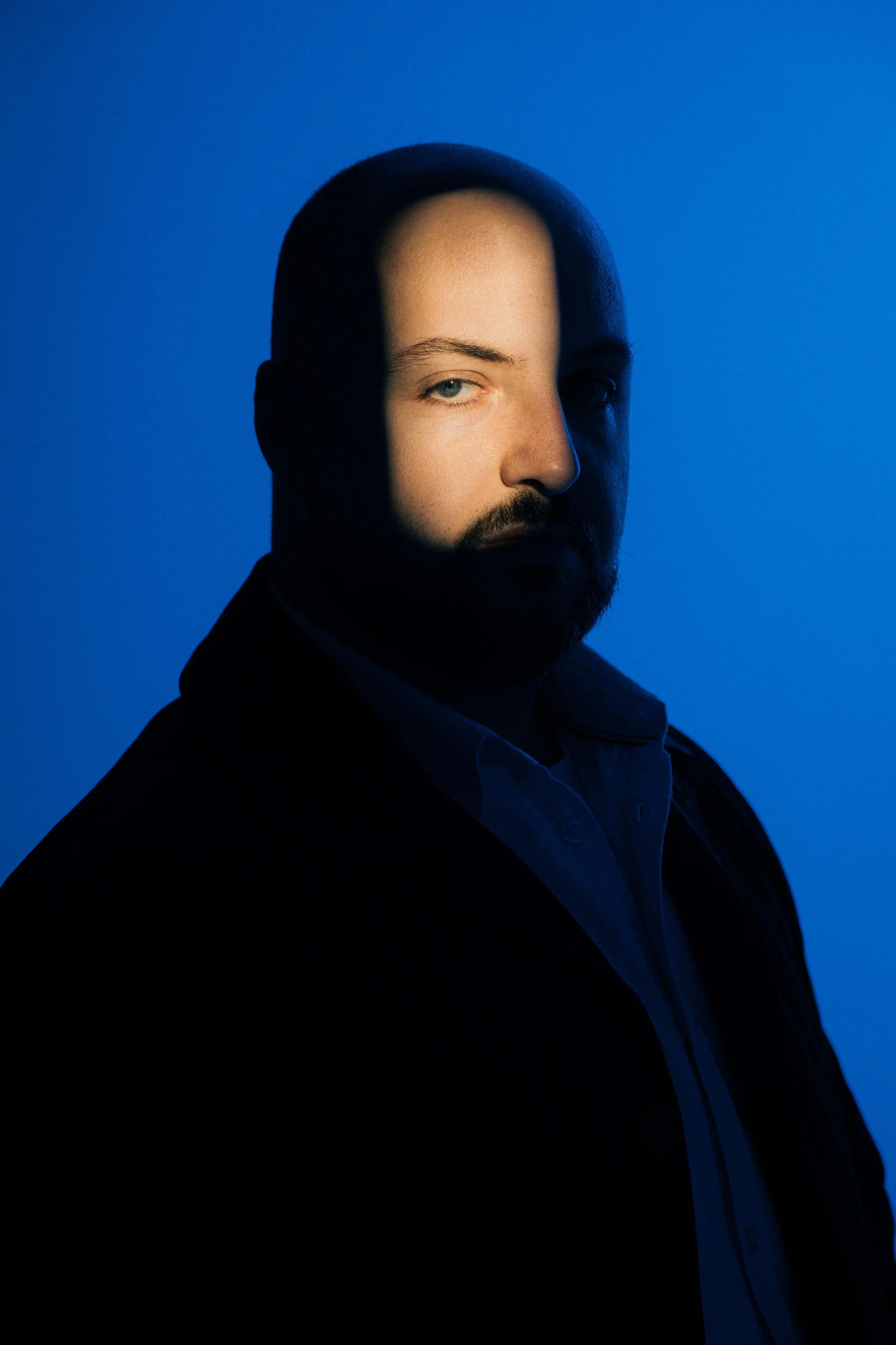 A portrait of a man with a beard and short dark hair, looking at the camera against a blue background, with a shadow cast across half of his face.