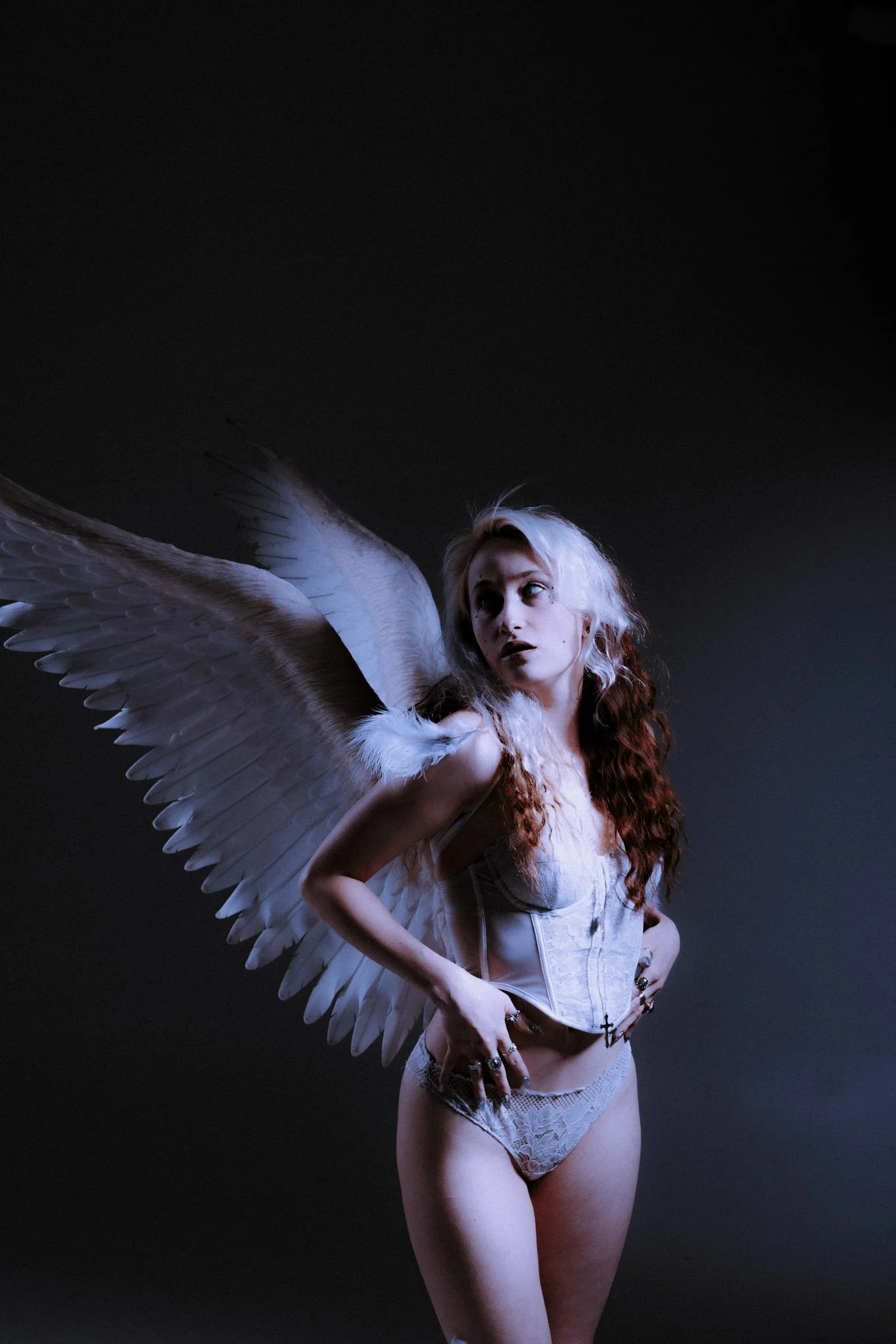 A woman with long, curly reddish-brown hair and light skin is wearing white lace lingerie and has large white angel wings. She is posing with her hands on her hips against a dark background.