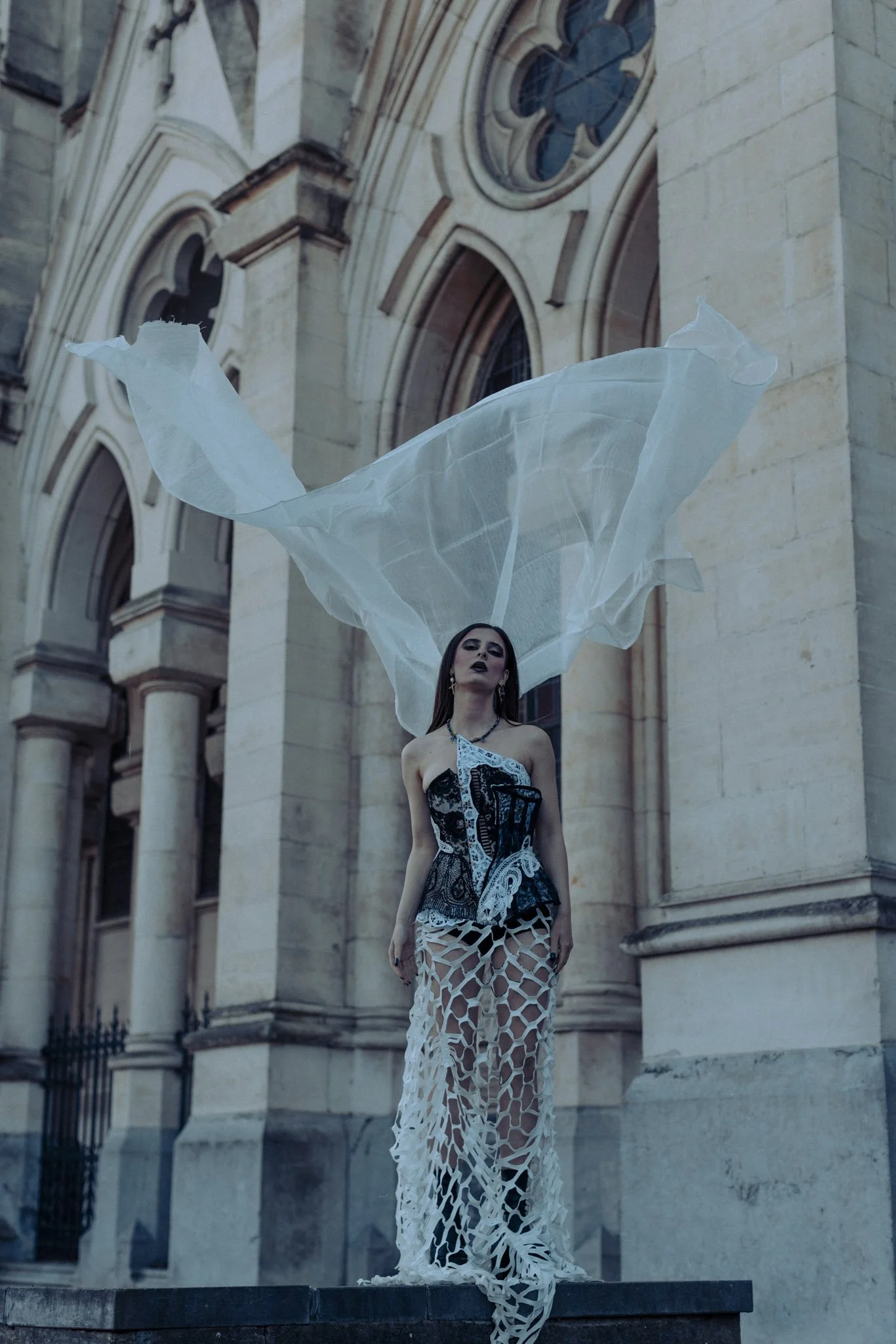A woman wearing a black and white dress with cut-out patterns stands in front of a historic stone building with stained glass windows, with flowing white fabric above her head.