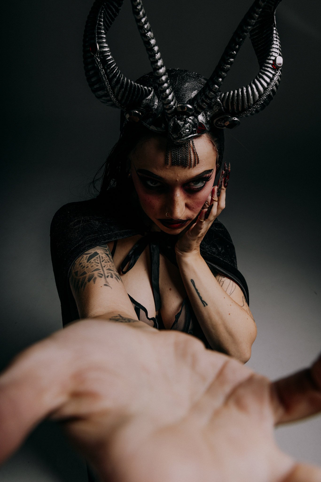 A woman with dark hair and tattoos on her arm, wearing a headdress with large black and white horns, touching her face with her hand, with intense makeup and a dark background.