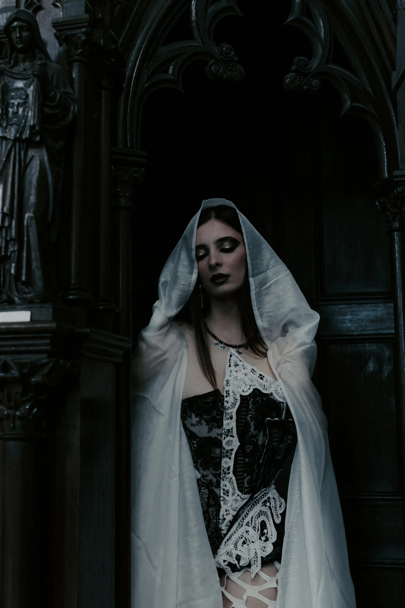 A woman with dark makeup and dark hair, wearing a gray veil, a black lace top, a white embroidered jacket, and fishnet stockings, standing in front of a dark, ornate gothic background with statues and elaborate woodwork.