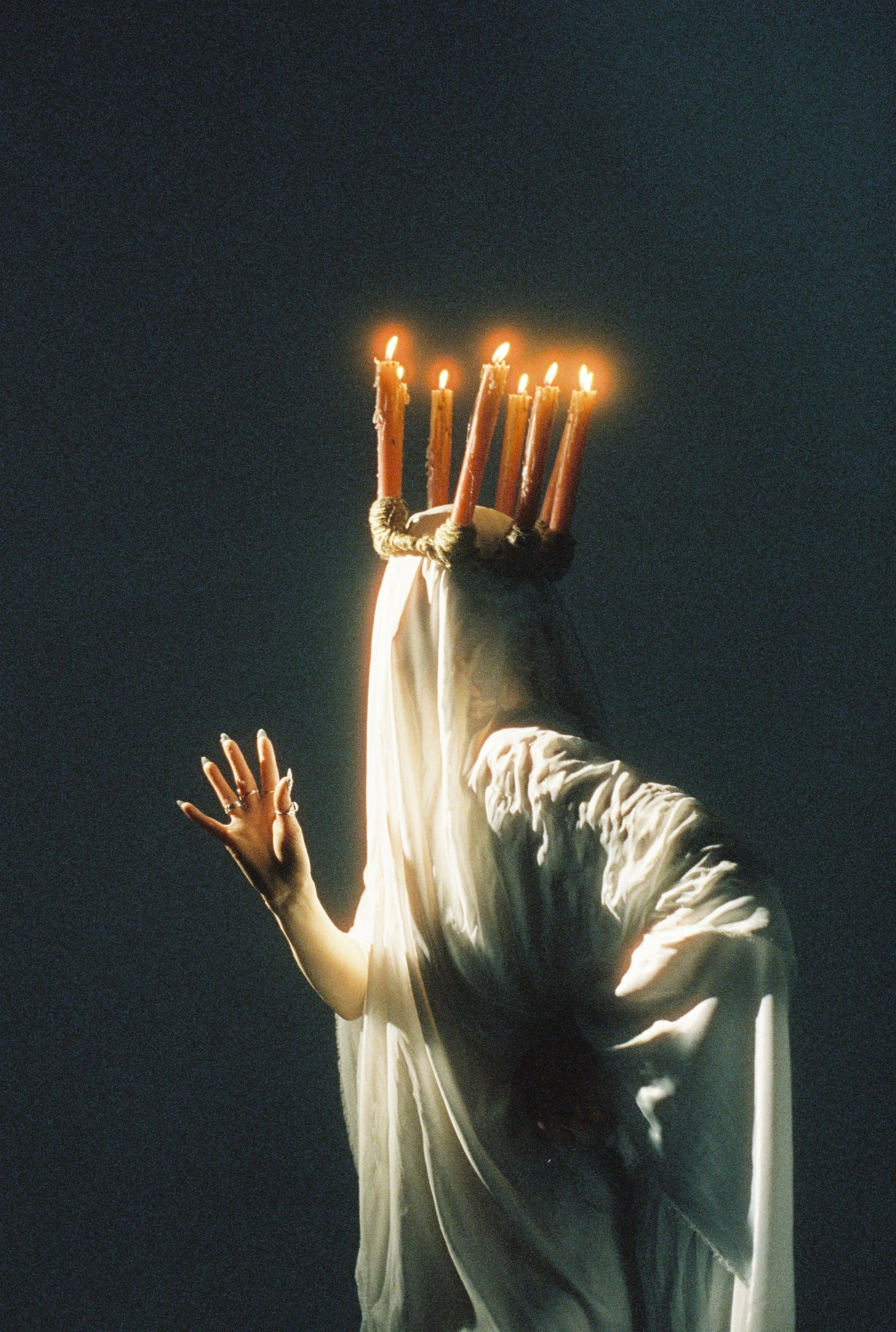 A person dressed in a white robe holding seven lit candles with wax dripping down, against a dark background, with an outstretched hand displaying rings.