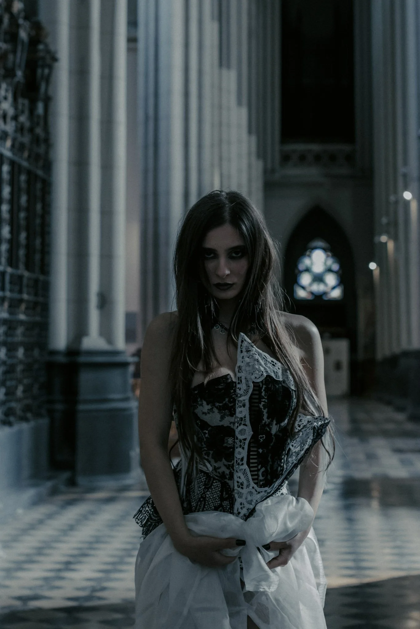 A young woman with long hair and dark makeup standing in a gothic cathedral with tall columns, stained glass windows, and a reflective water floor.