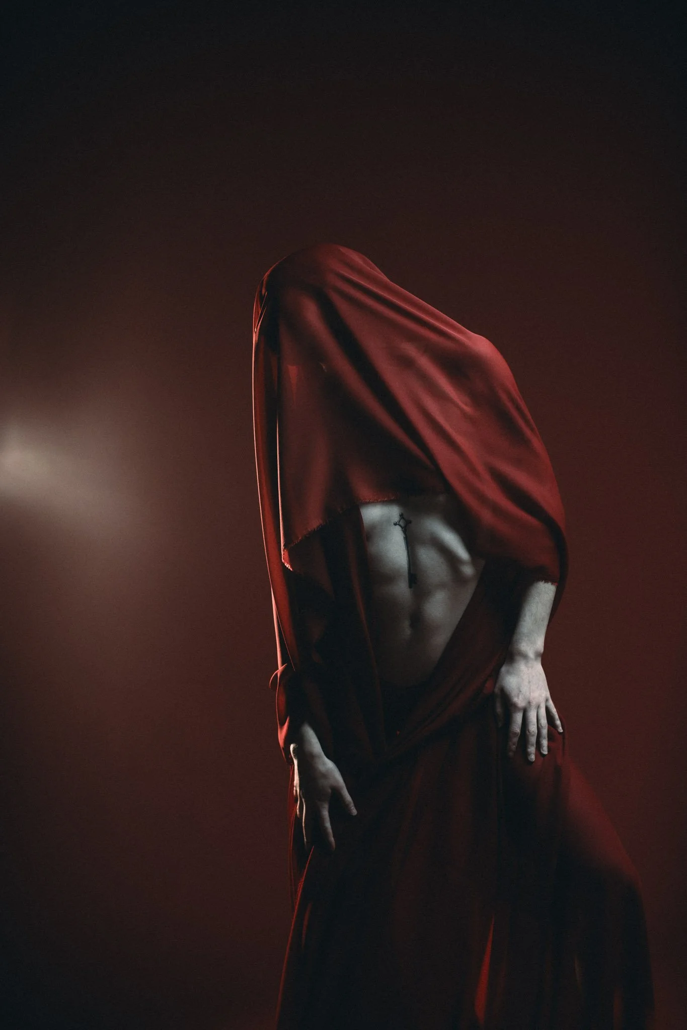 Person with red and black draped fabric covering their head and face, partially revealing their torso and arm, set against a dark background with red lighting.