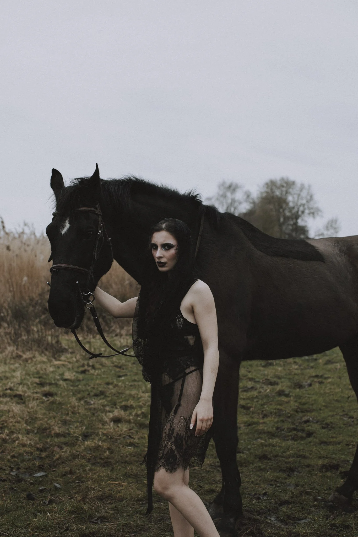 A woman with black hair and dramatic makeup standing next to a black horse in an open field during dusk.