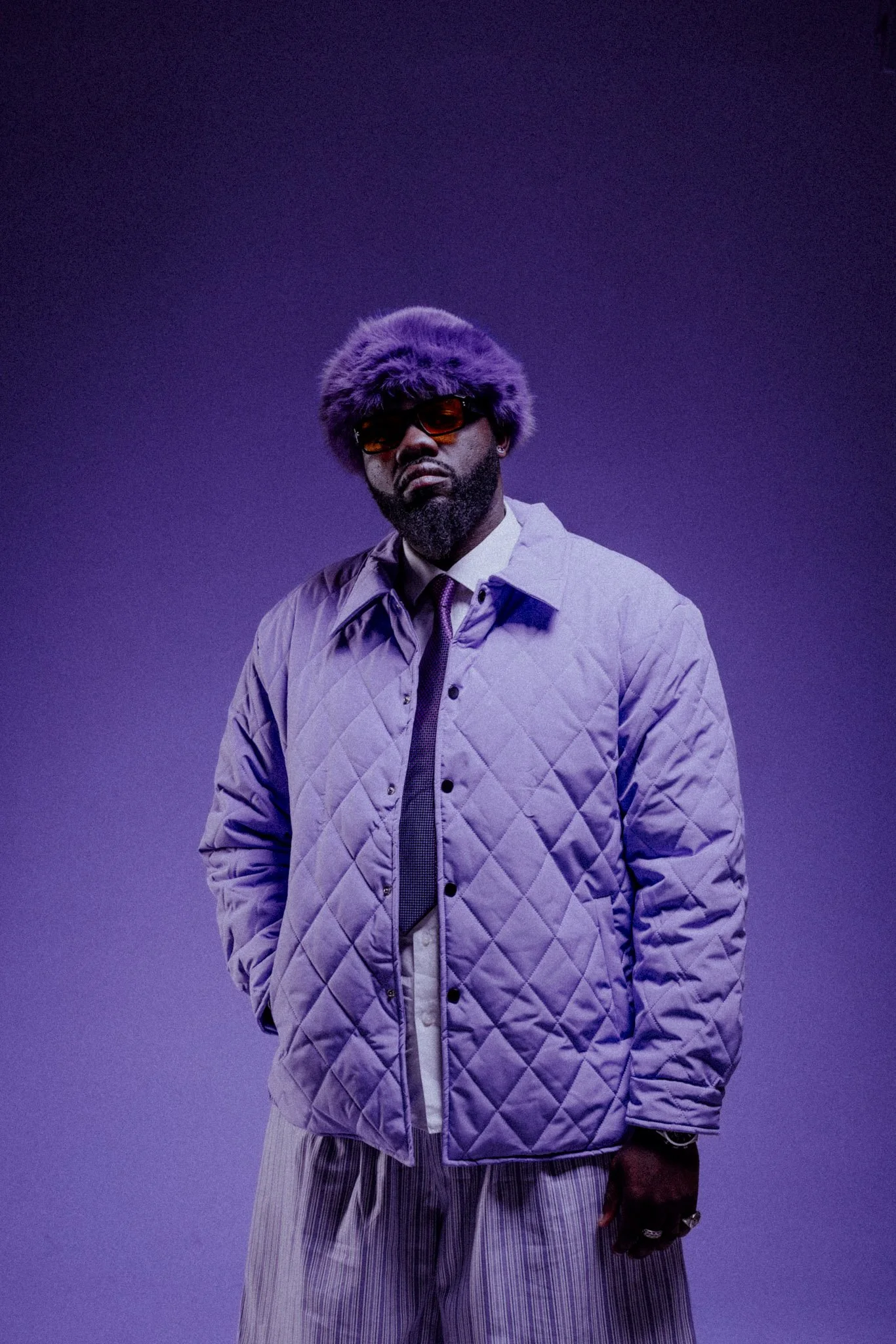 A man wearing a purple quilted jacket, striped pants, with a large furry purple hat, sunglasses, and gloves, standing against a purple gradient background.