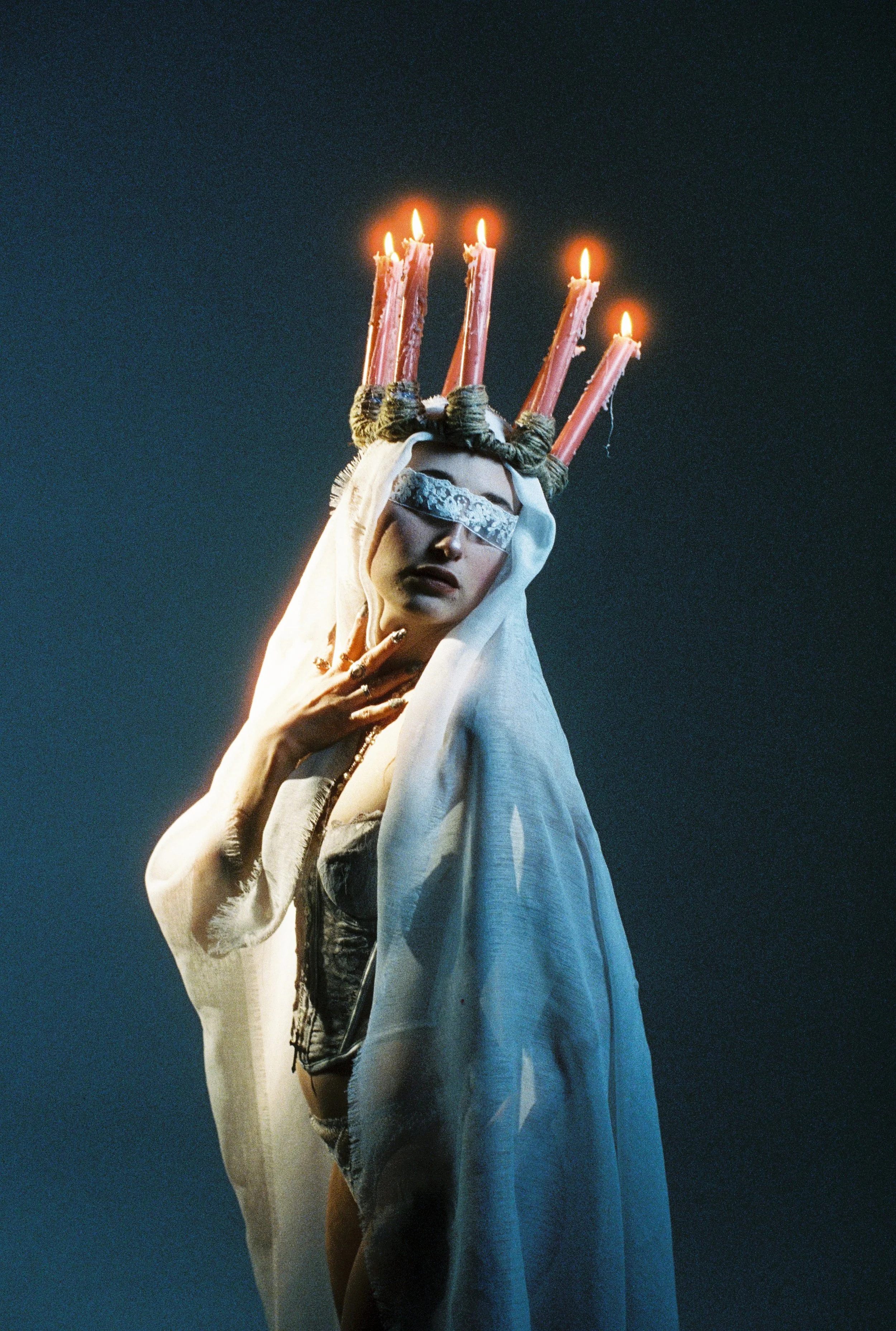 A person with long hair and jewelry, dressed in a white garment, wearing a crown made of candles, with a dark background.