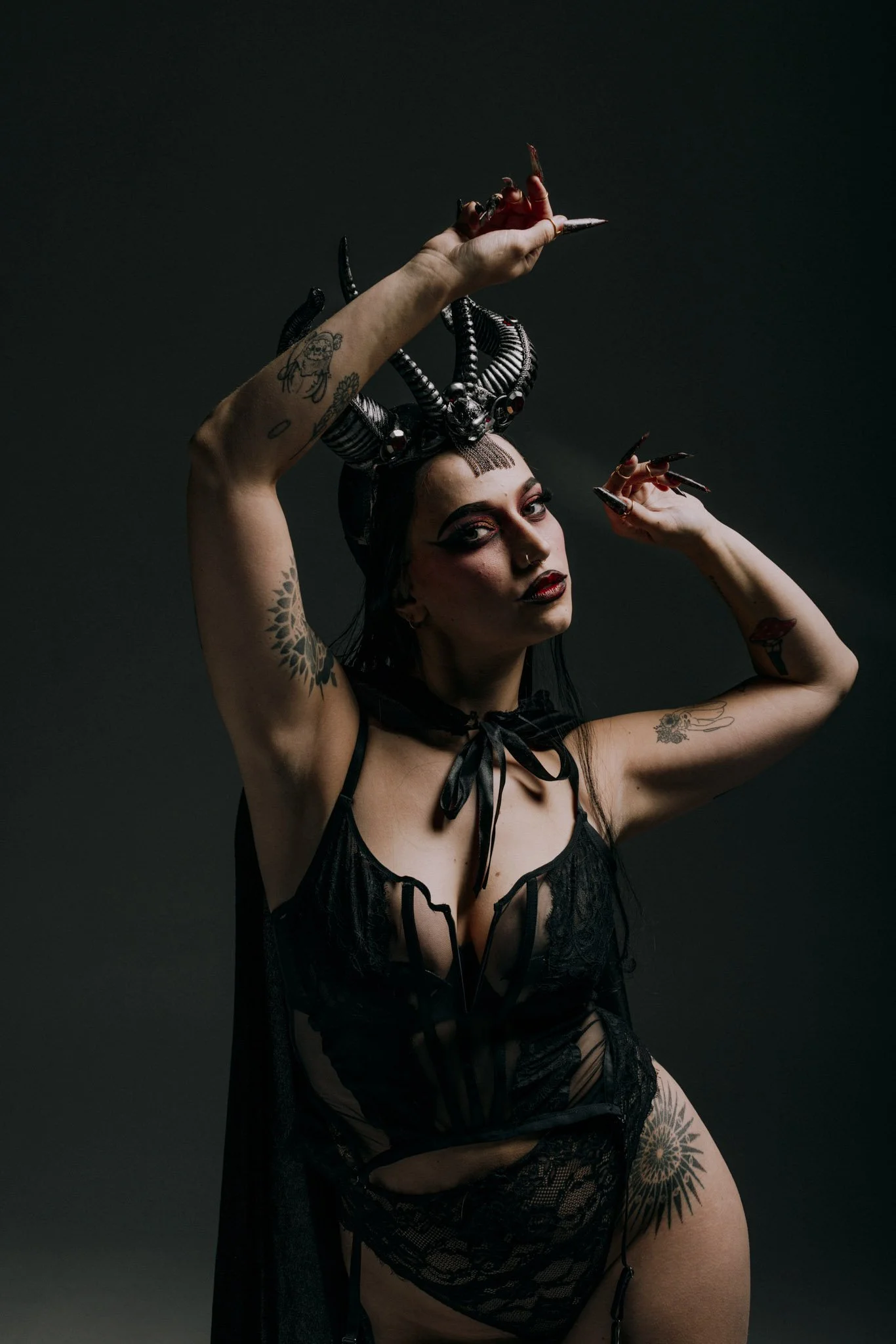 A woman with tattoos and dark makeup posing against a dark background, wearing black lace lingerie and an elaborate black snake headdress.