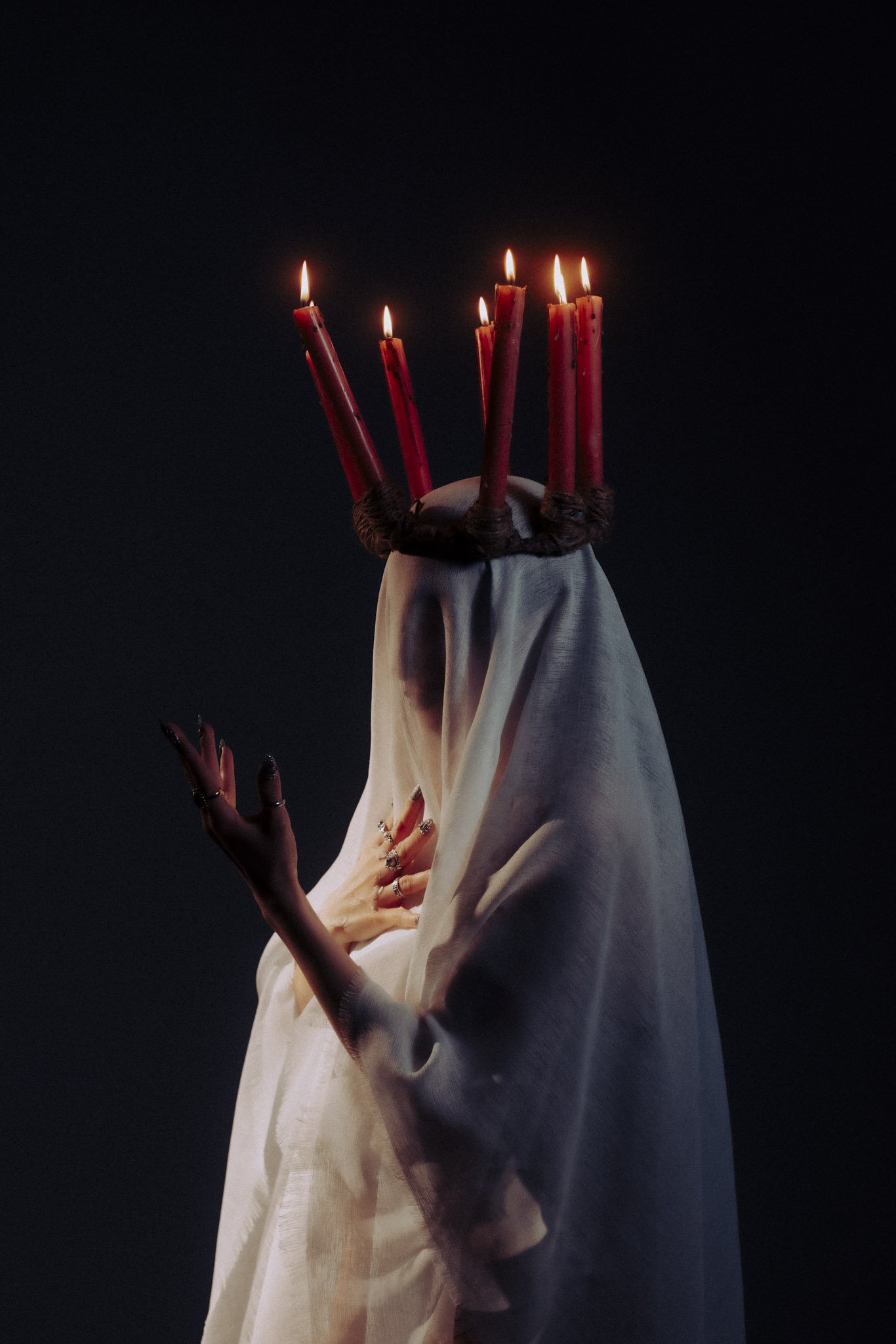 A figure with a white flowing garment and multiple red candles on their head, with flames lit, against a dark background.