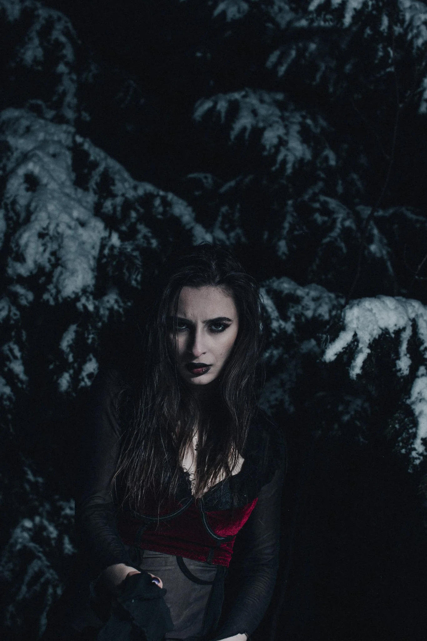 A woman with dark hair and intense makeup, dressed in black and red clothing, standing in a dark, snowy forest at night.