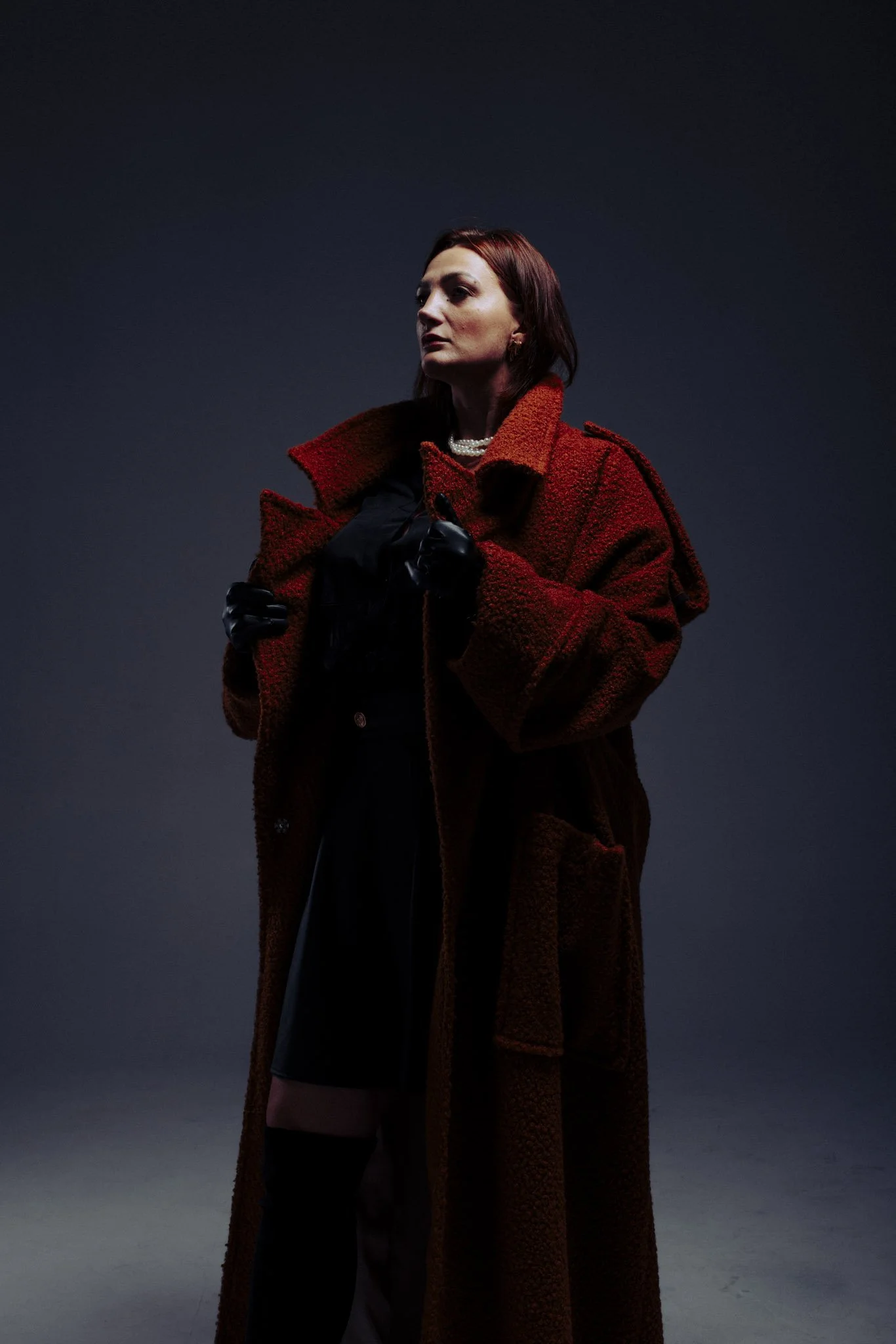 A woman with red hair wearing a red coat and black gloves, standing against a dark background with a serious expression.