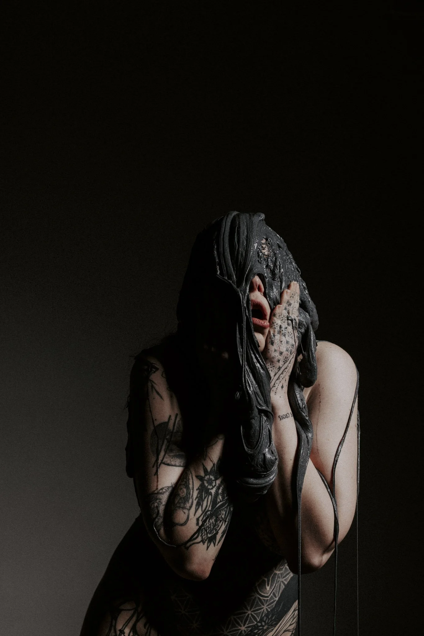 A person with tattoos covering their arm and chest is holding their head in their hands, covering their face, with long black hair hanging over their face. The background is black and the lighting is focused on the person.