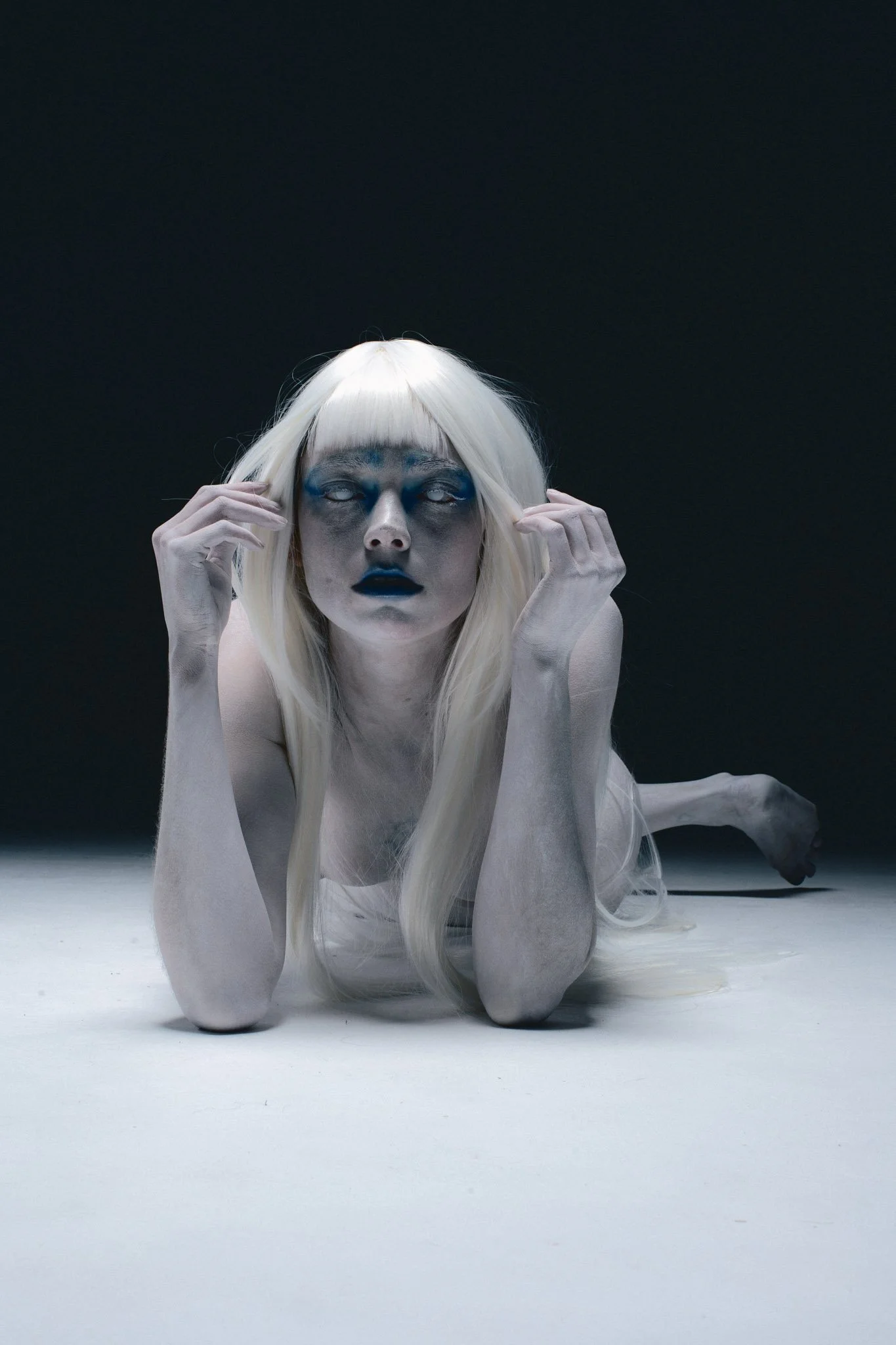 A person with long platinum blonde hair is lying on a white surface with a dark background. They have artistic makeup with blue and black paint around their eyes and dark lipstick. The person is holding their face with both hands, looking directly fo