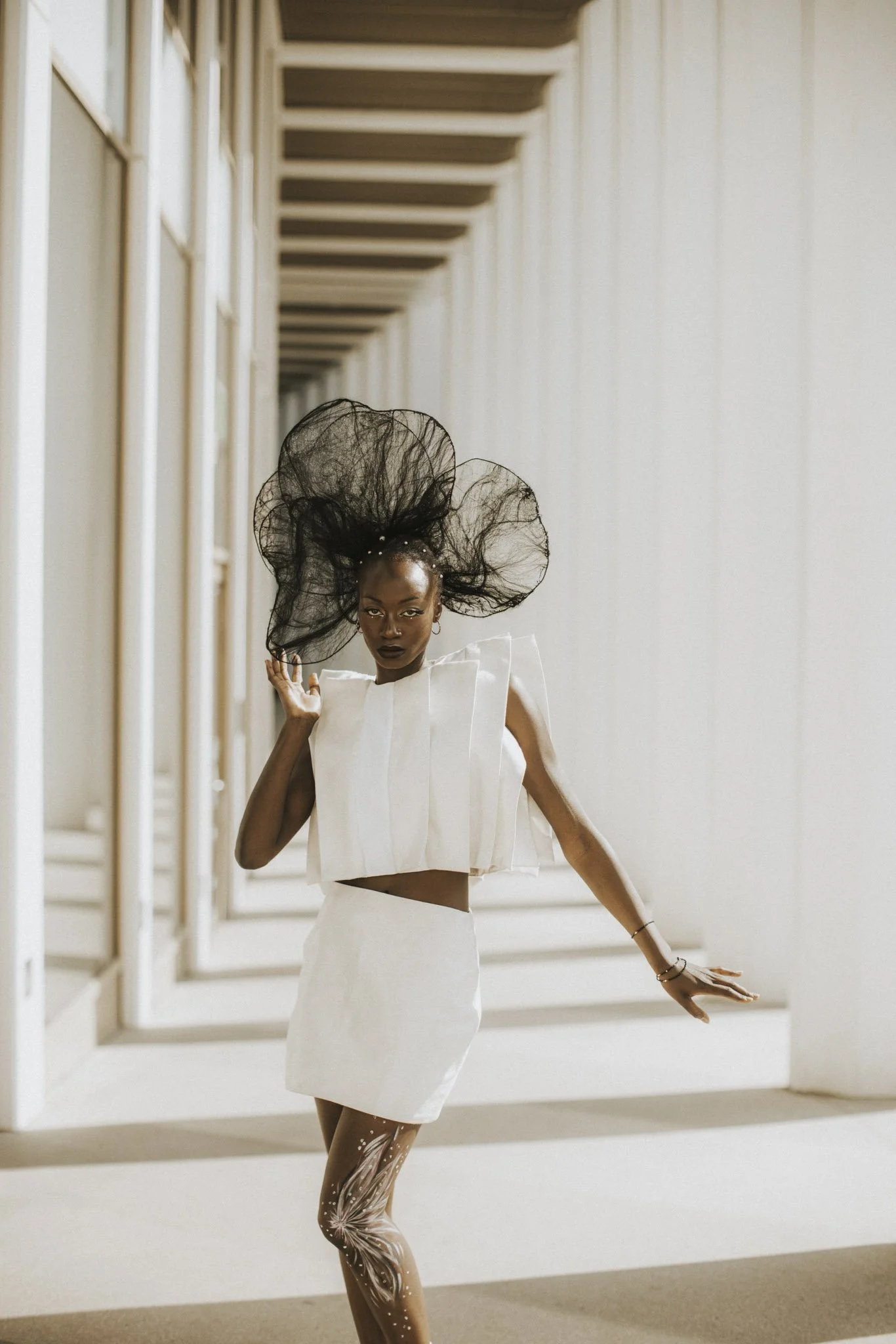 A woman wearing a white outfit with unique design and a large black, netted headpiece, standing in a modern, minimalist corridor with white walls and vertical beams.