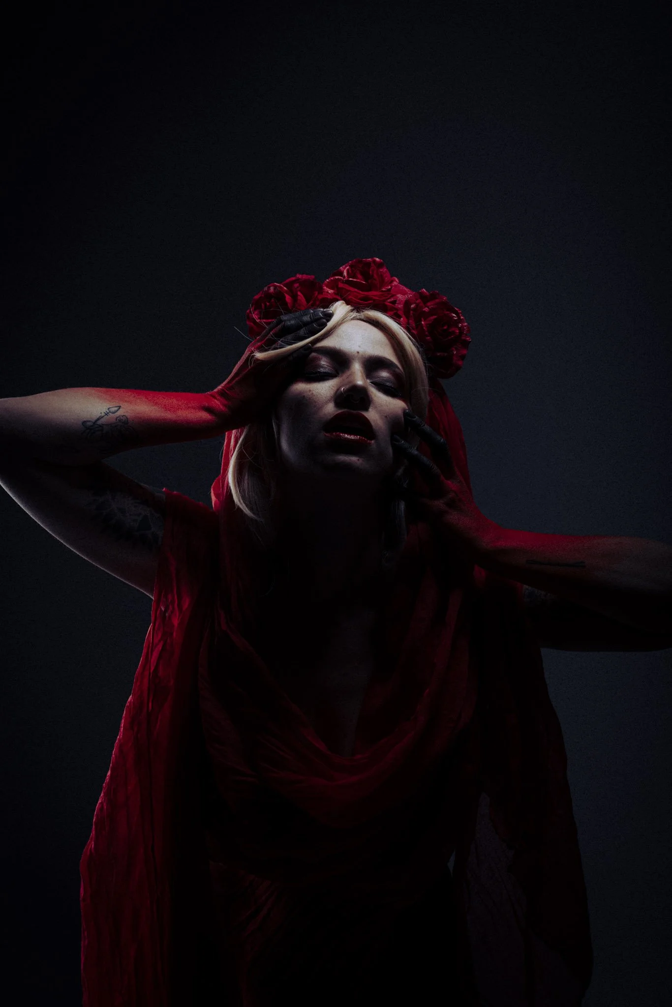 Woman with blond hair, wearing a red headpiece made of roses, red dress, and black gloves, posing with her head tilted back and eyes closed against a dark background.