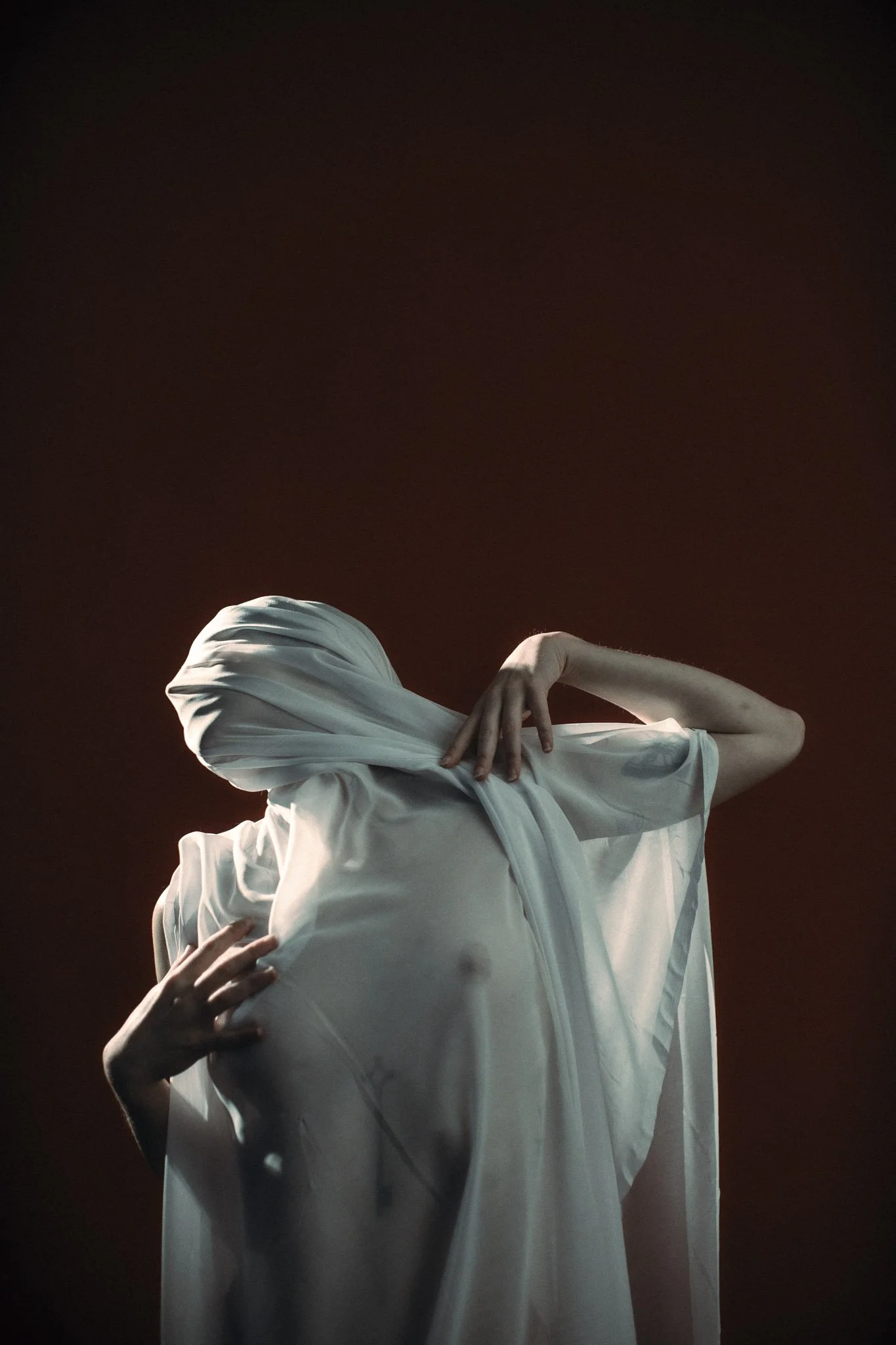 Person wearing a white shirt, standing against a dark background, with arms crossed over the chest and head turned away.