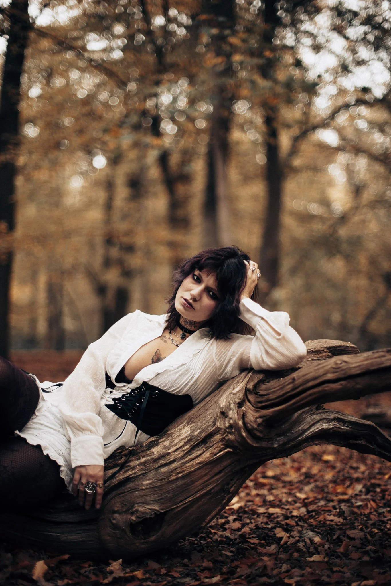 A woman with dark, wavy hair and tattoos, dressed in a white blouse, is lying on a large fallen tree in a forest during autumn. She has a serious expression and is touching her head with one hand, surrounded by autumn leaves and blurred trees in the 