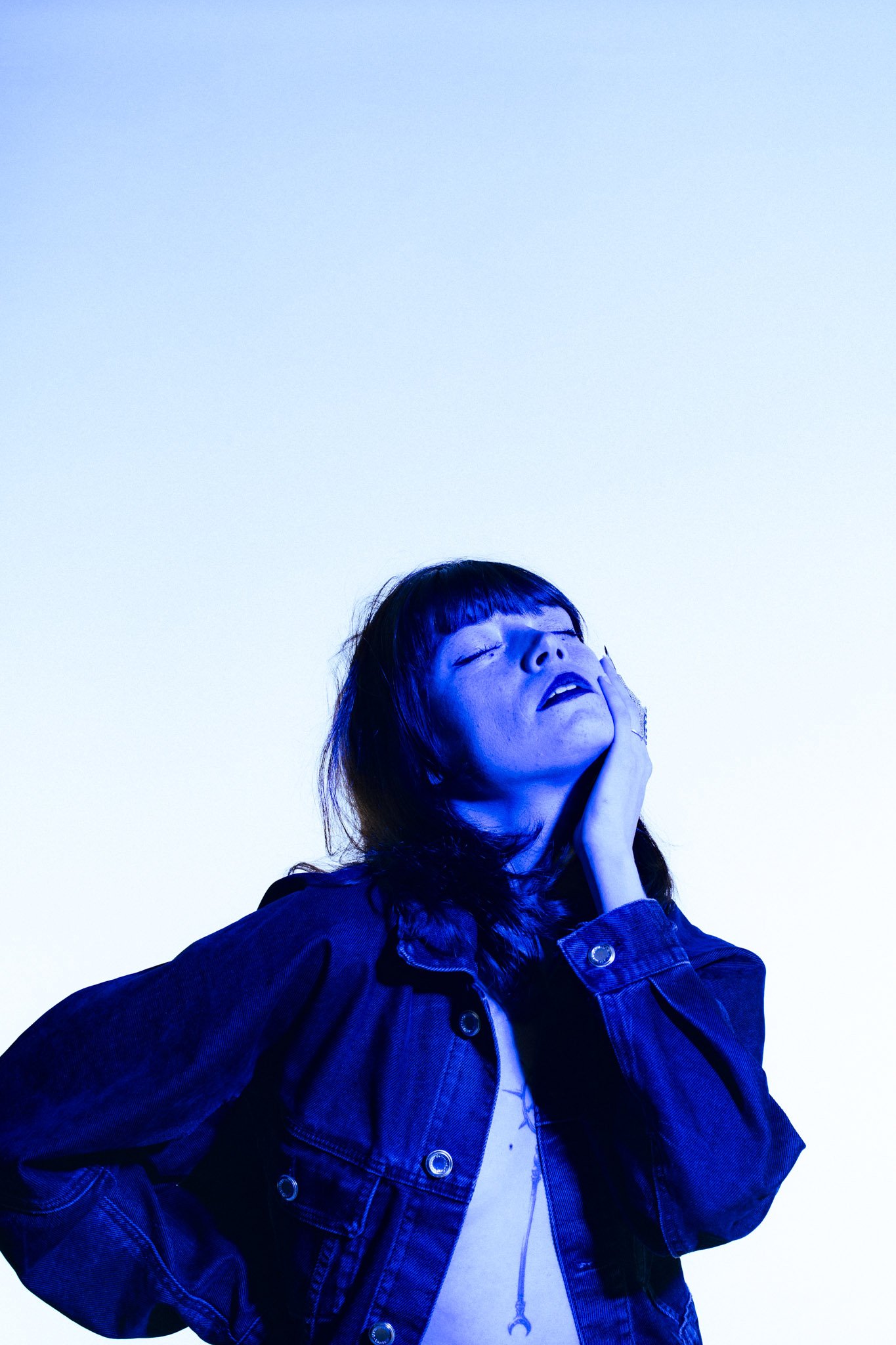 A woman with dark hair, bangs, glasses, and a denim jacket poses with her eyes closed, touching her face, against a gradient blue background.