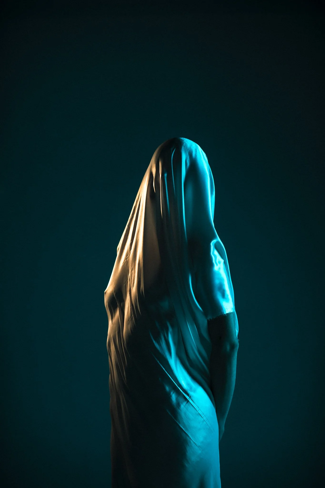 A person is covered with a white sheet, creating the appearance of a ghostly figure against a dark background, illuminated with blue and beige lighting.