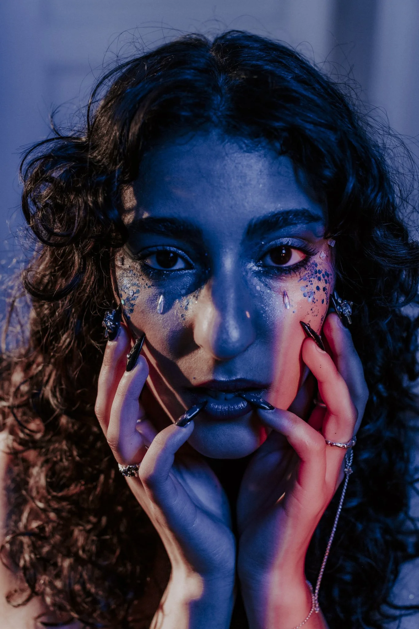 A close-up of a woman with curly hair and colorful makeup, holding her face with her hands, featuring multiple rings and dark nail polish, with dramatic lighting and a dark background.