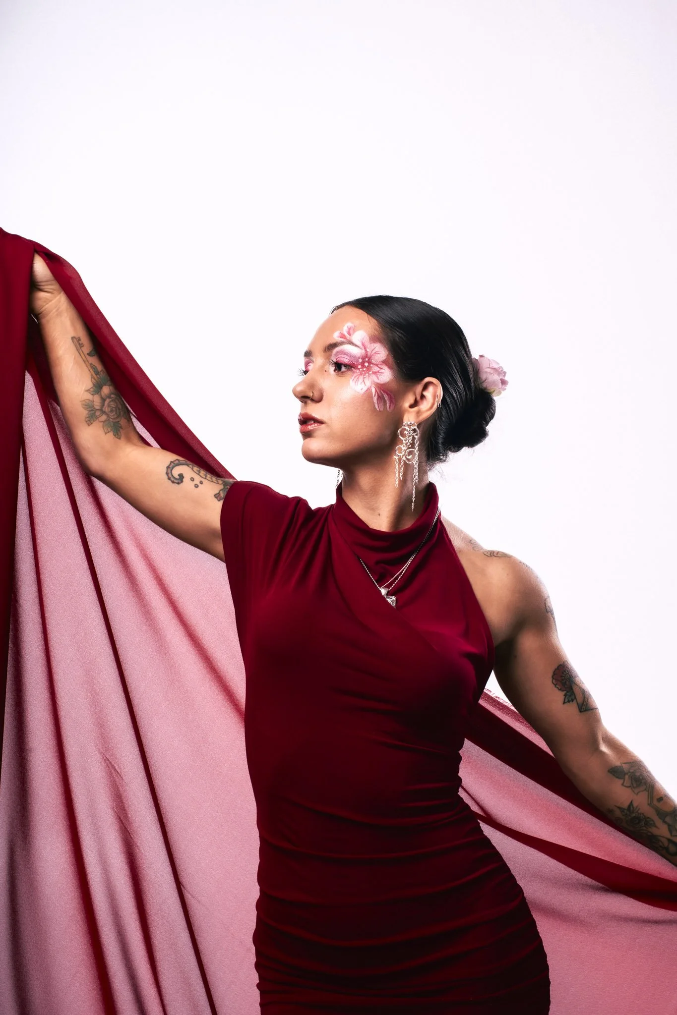 A woman with black hair styled in a bun, decorated with a pink flower, is adjusting pink fabric with her arm raised. She has floral face makeup, earrings, and tattoos on her arm. She is wearing a red sleeveless dress and a necklace with a pendant, po