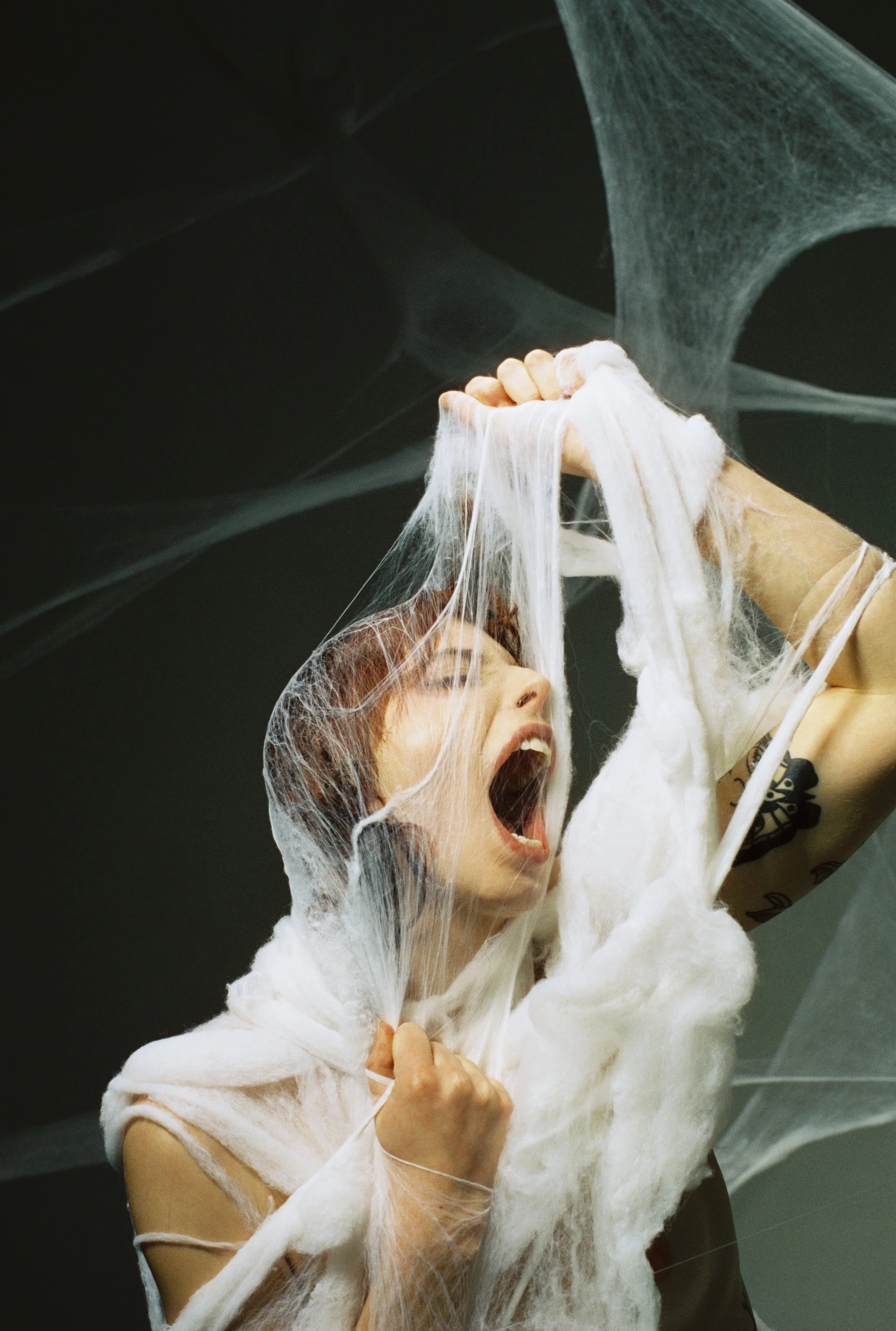 A person with short hair, wearing a beige shirt, is passionately singing or shouting while holding a handful of white cotton in a dark room with spools of white thread or cotton webs in the background.