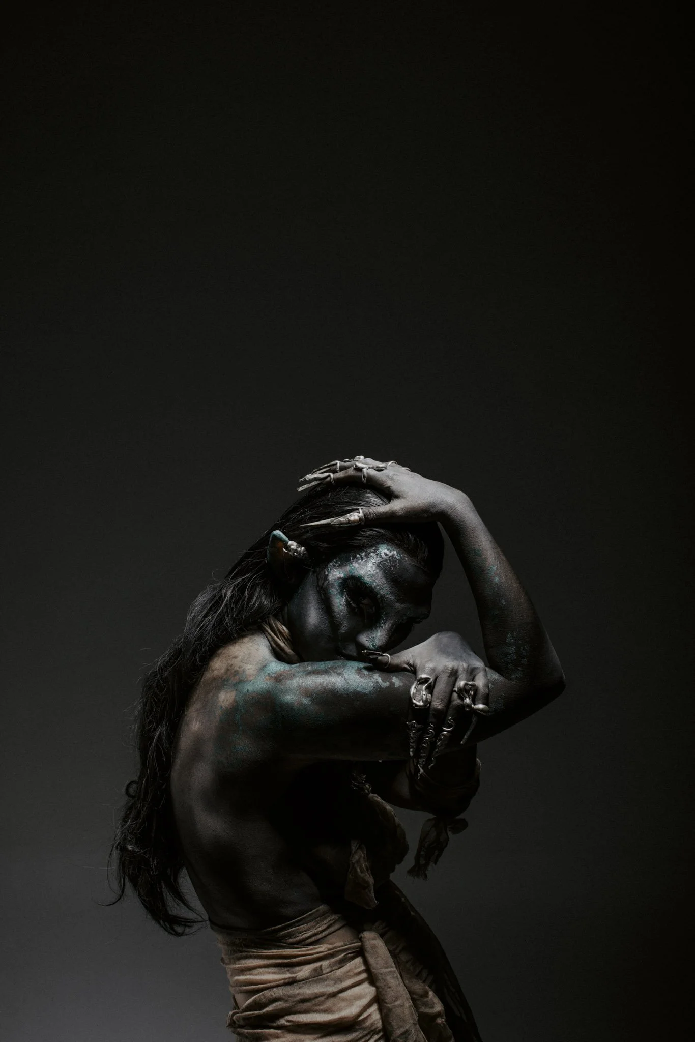 A woman with painted face and body, holding her head and arm, dressed in earthy tones and jewelry, standing against a dark background.