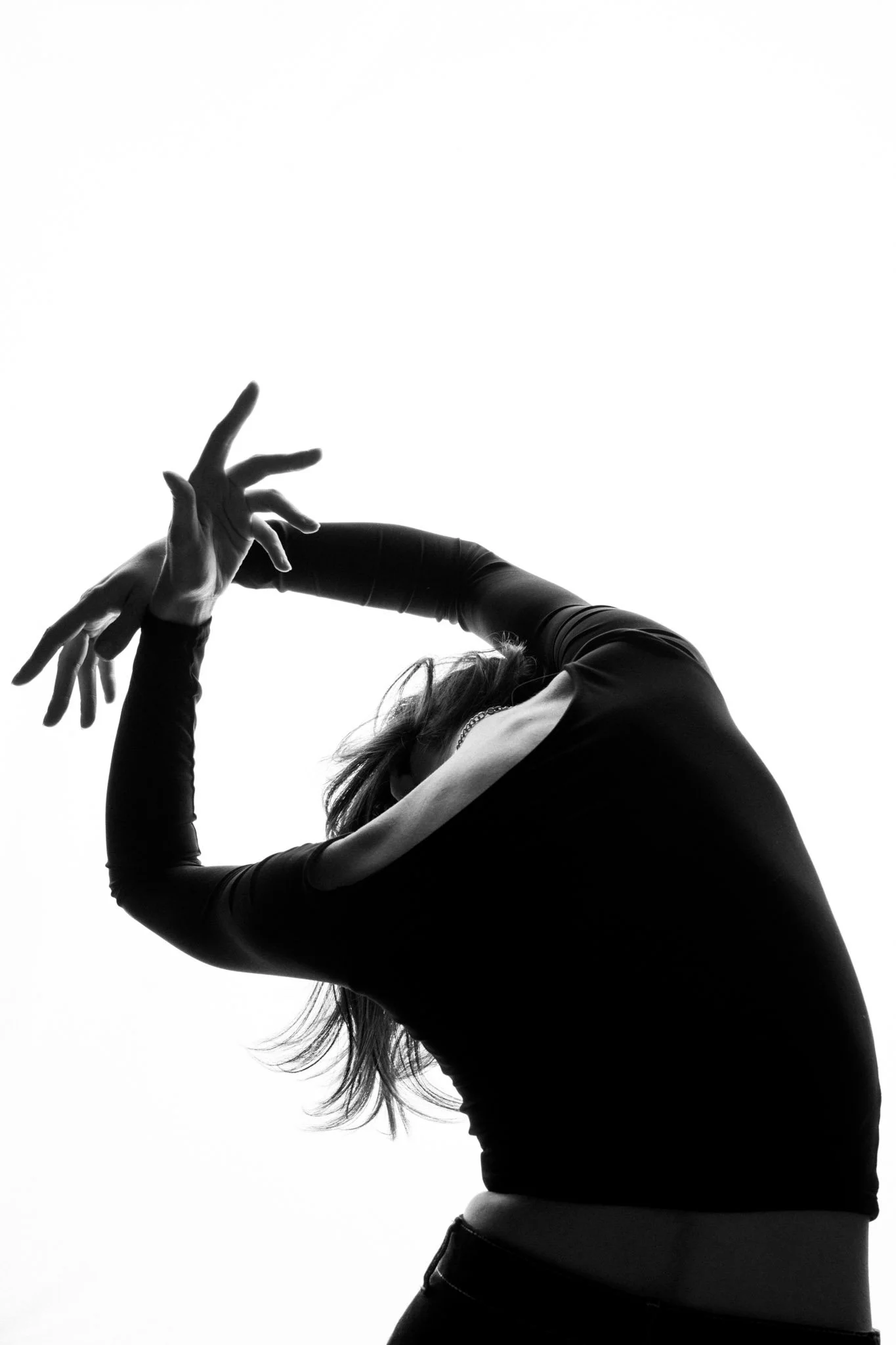 A black and white photo of a person with long hair doing a dance pose with their arms raised and hands near their face, wearing a long-sleeve top and high-waisted bottom, against a plain white background.