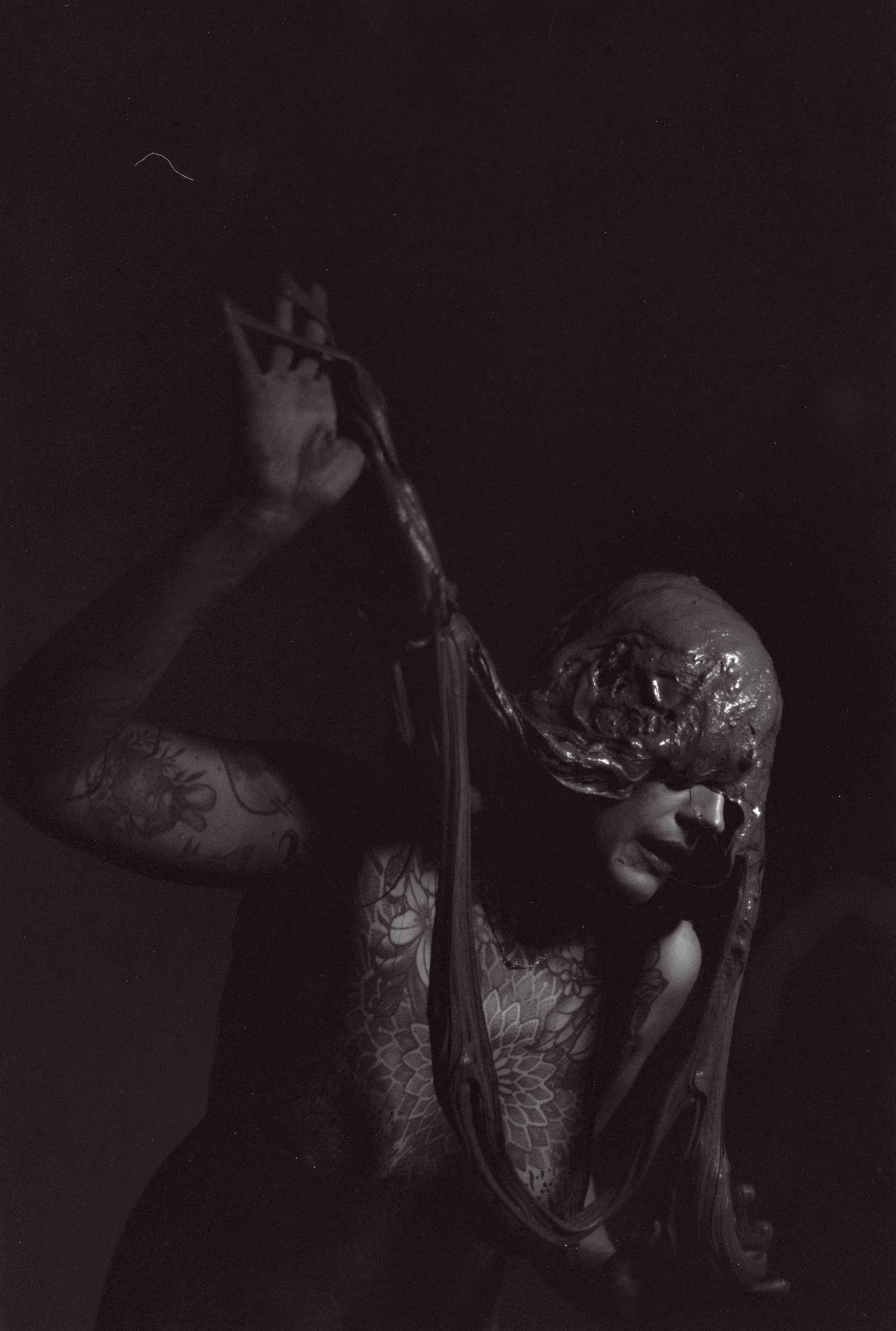 A woman with tattoos and laser hair growth on her face is holding a small sculpture of a head with curly hair and an open mouth. The sculpture appears to be metallic and detailed.