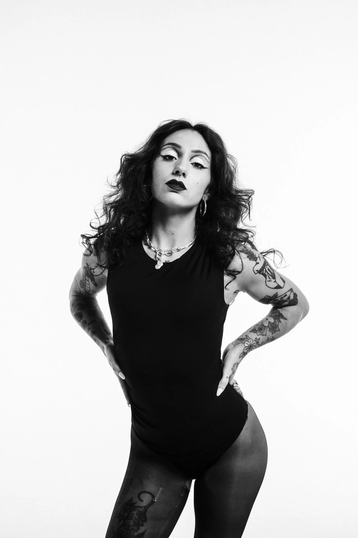 Black and white photo of a woman with dark, curly hair and tattoos on her arms and thigh, wearing a sleeveless dark outfit, with a confident pose and serious expression.