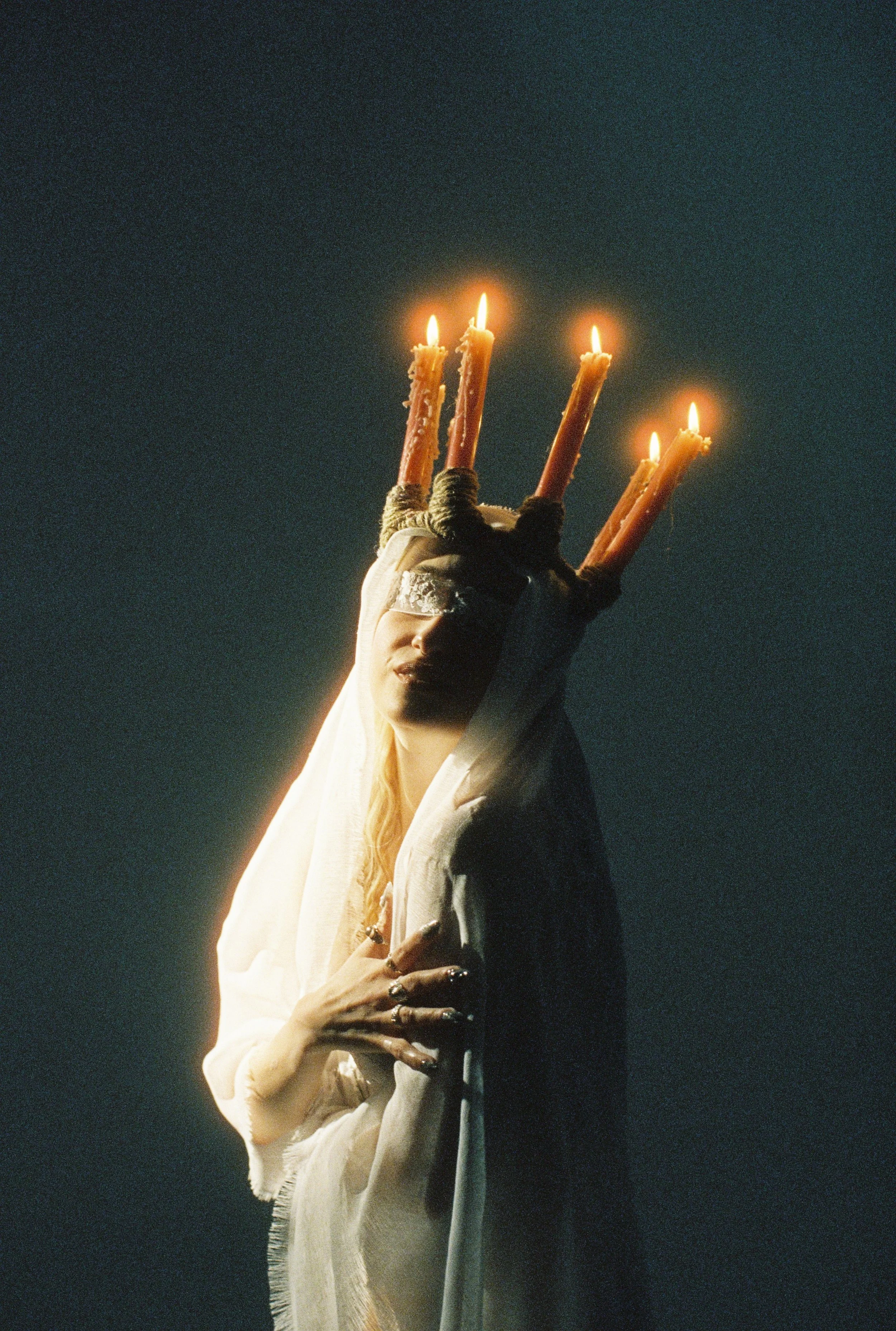 A person holding a hand with four lit candles tied with rope, dressed in a white garment, against a dark background.