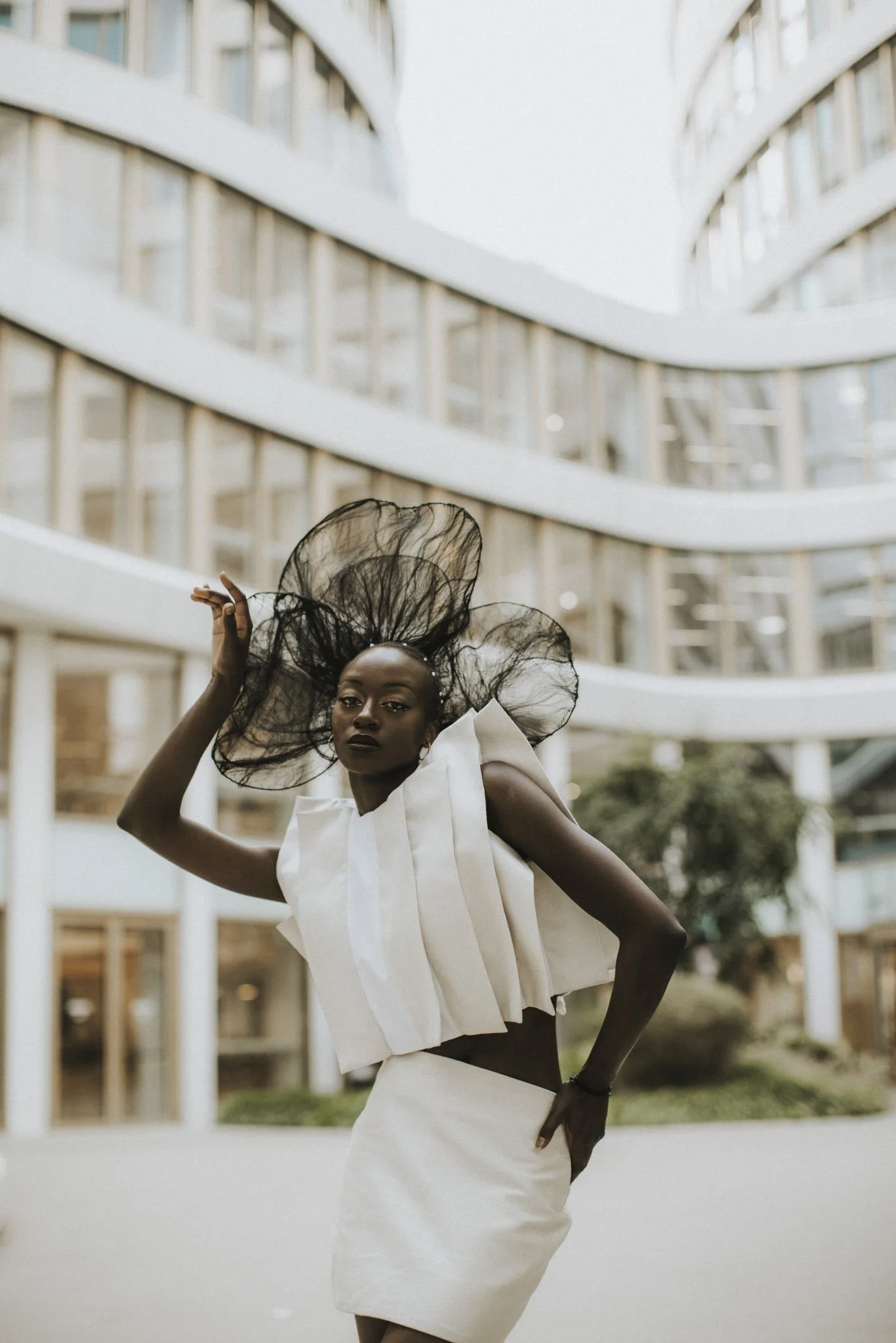 A Black woman with elaborate black hair and dark lipstick is posing outdoors in front of a modern building with curved white balconies. She is wearing a white, asymmetrical fashion ensemble with large ruffle details, and has one hand on her hip with 