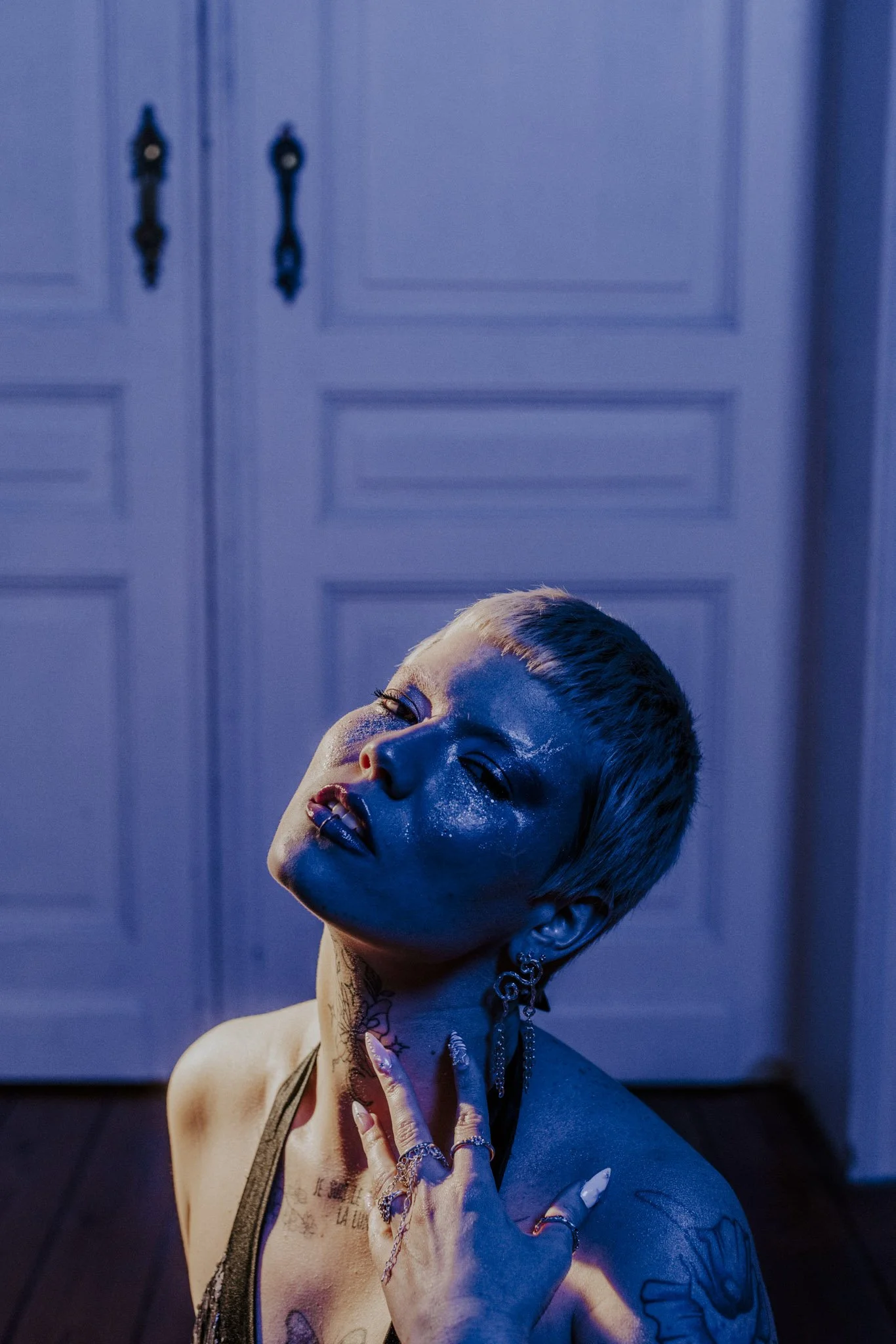 A person with short hair and tattoos poses with eyes closed and head tilted back, illuminated by blue and purple lighting in front of a paneled wall.