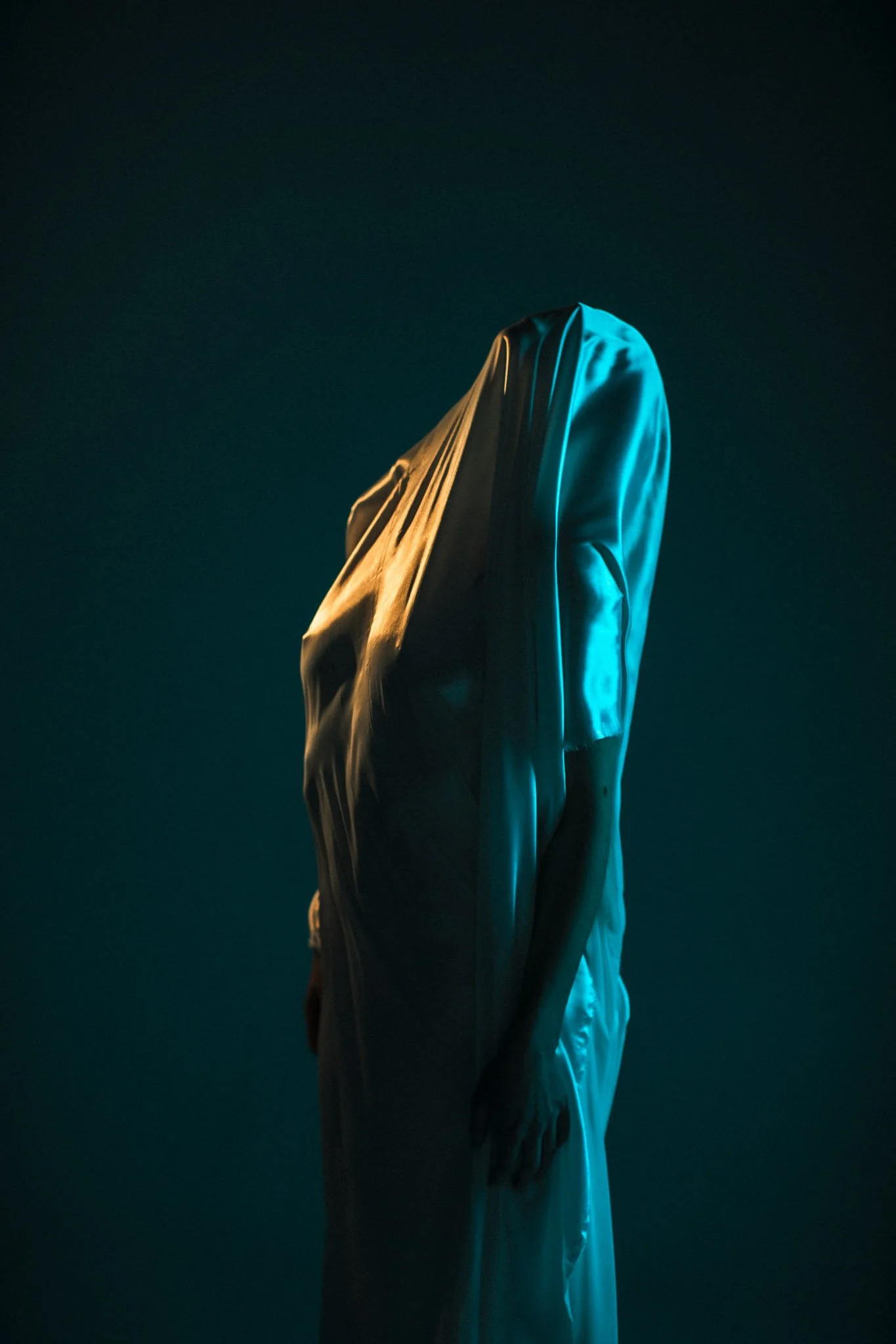 A person standing with a cloth draped over their head and shoulders, illuminated by blue and yellow lighting against a dark background.