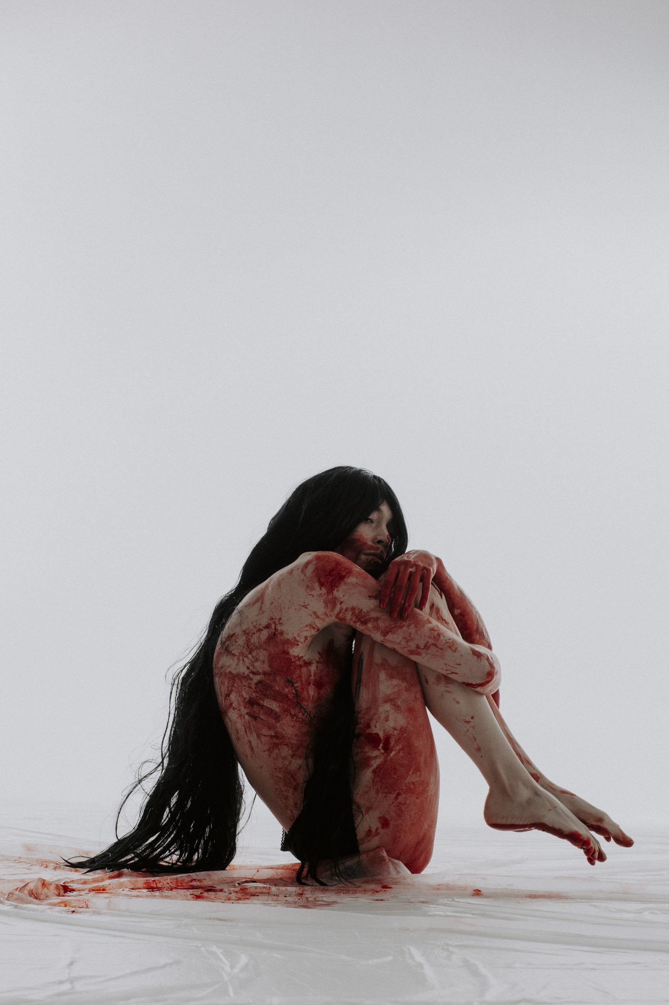 A woman with long black hair, covered in red streaks, is sitting on the ground with her arms wrapped around her knees, against a plain white background.