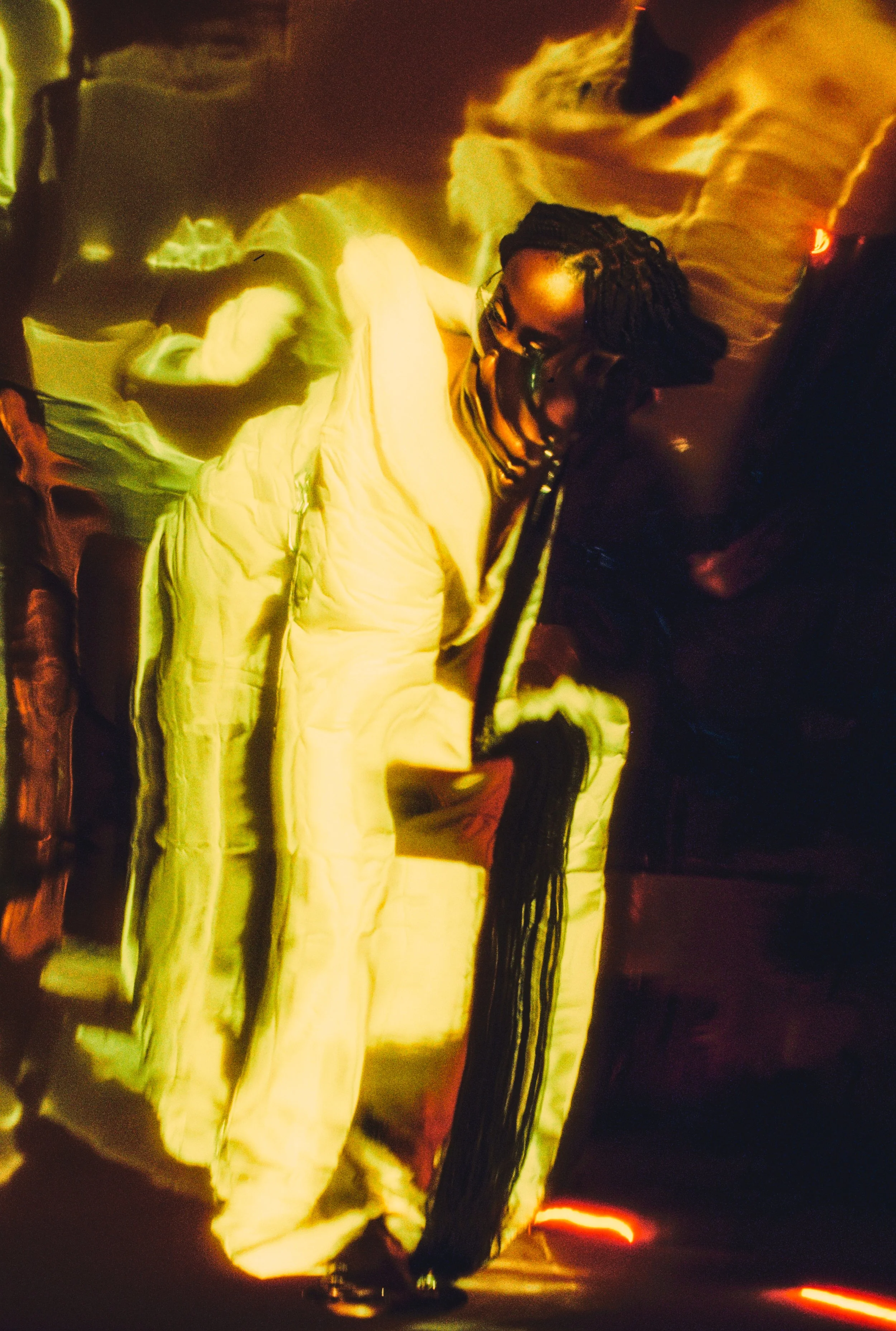 A person with glasses and long braided hair leaning against a reflective surface in a dimly lit environment with warm orange and yellow lighting.
