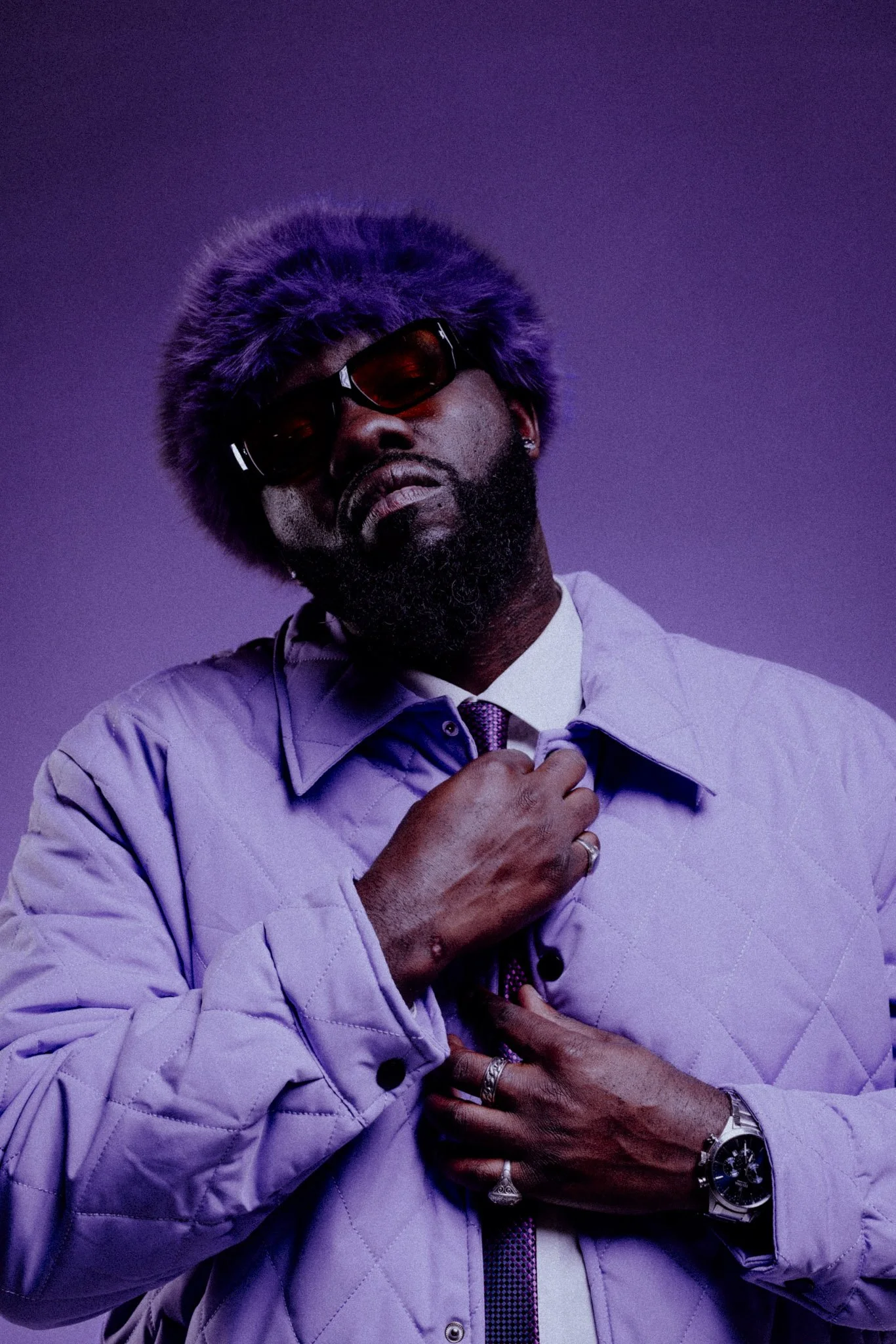 A man with a beard wearing dark sunglasses, a purple furry hat, a light purple quilted jacket, and a dress shirt with a tie, adjusting his collar. The background is purple.