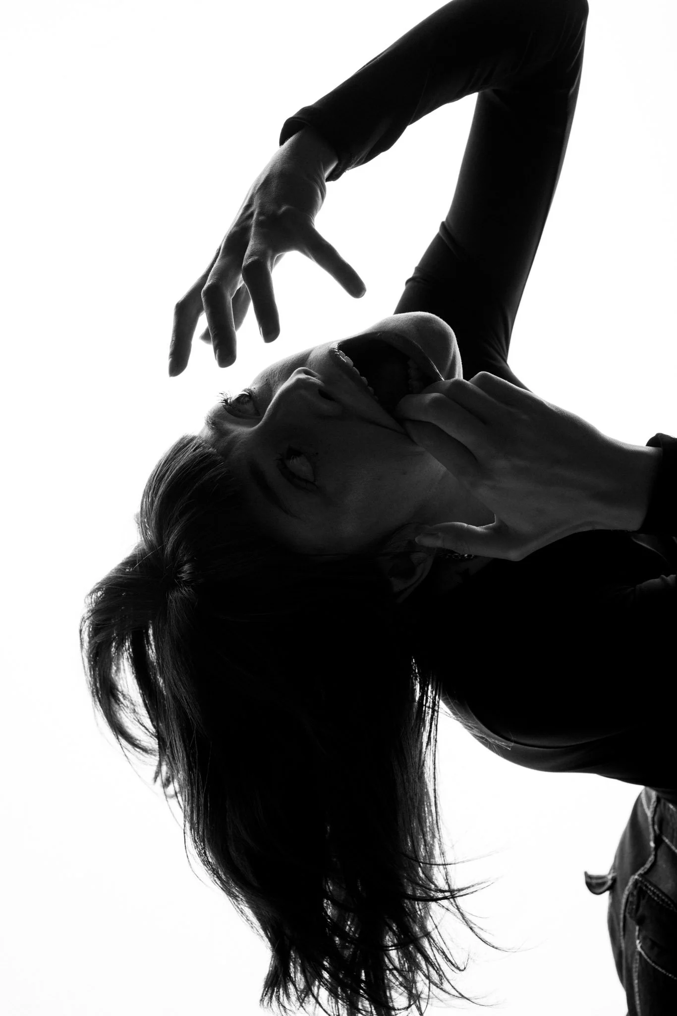 A black and white photo of a woman with long hair, posing with her head tilted back and her hands framing her face, creating a dramatic and artistic silhouette.